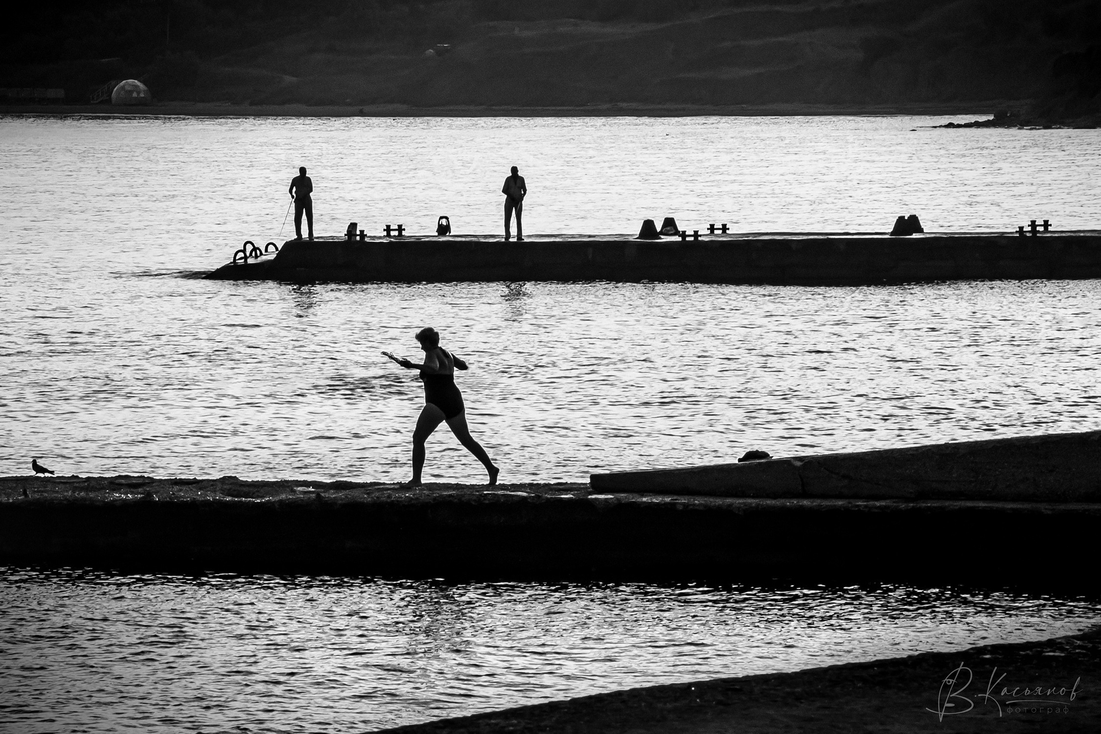 Женщина бежит по причалу чтобы нырнуть в воду, два мужчины наблюдают за ней