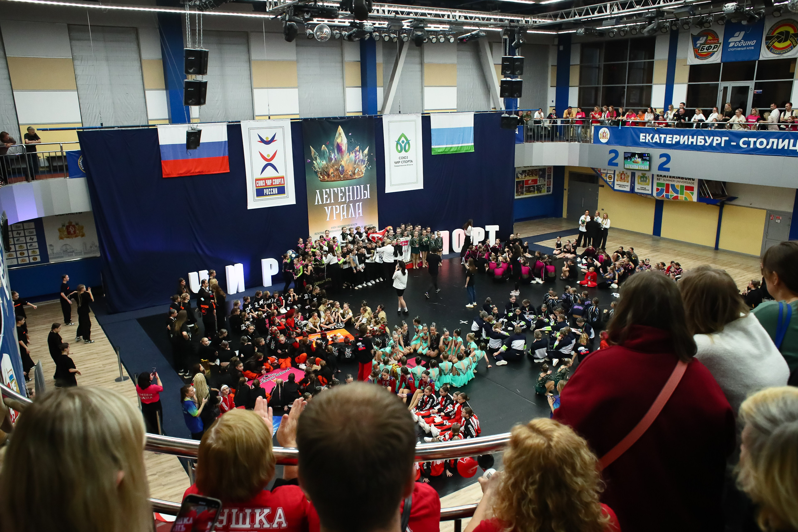 Общий вид спортивной площадки на которой проходит церемония награждения участников соревнований по чир-спорту. На переднем плане головы зрителей наблюдающих за происходящим на площадке, далее сами участники, на заднем плане пьедестал на котором стоят победители, за ними расположены банеры и темно-синяя драпировка, по бокам буквы ЧИР и СПОРТ.