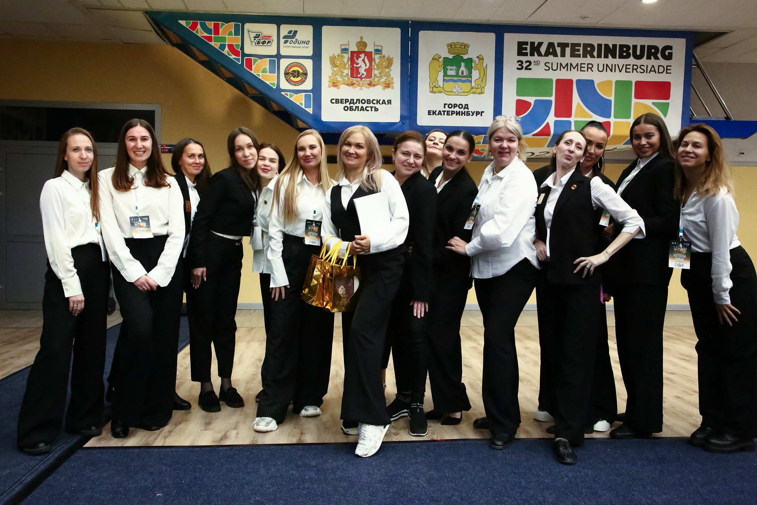 Группа из 14-ти судей соревнований по чир спорту. В руках у некоторых из них наградная продукция. Они одеты в строгую одежду черно-белых тонов.