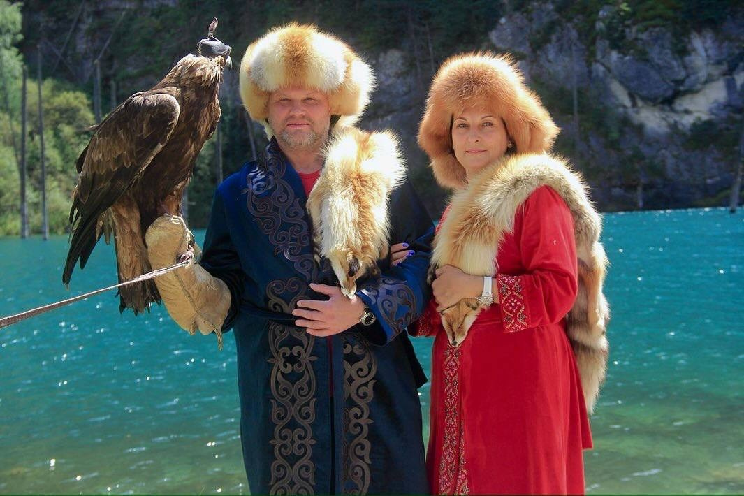 Мужчина и женщина в традиционных казахских костюмах позируют на фоне бирюзового озера Каинды. Мужчина одет в синее украшенное халатом, с меховой шапкой и держит орла на перчатке. Женщина в красном платье с меховой опушкой и шапке, украшенной мехом. Атмосфера традиционности и природной красоты.