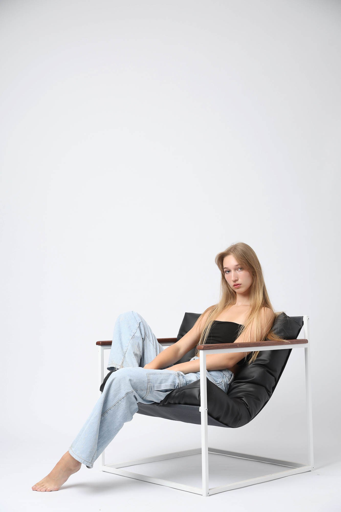 Модель в светлых джинсах сидит в кресле на белом фоне