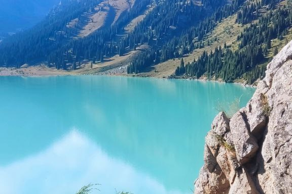 Величественный Чарынский каньон в районе Алматы с ярко-красными скалами, пересечённый извилистой дорогой, на которую падают длинные вечерние тени. Захватывающее сочетание красочных утёсов, зелёных склонов и драматичного неба создаёт эффектный пейзаж