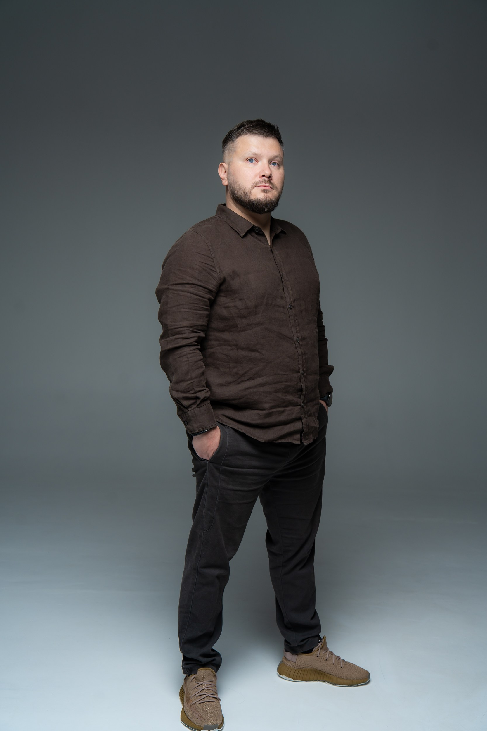 Мужчина в темной одежде на фоне белой циклорамы в студии, руки в карманах, уверенный взгляд в камеру