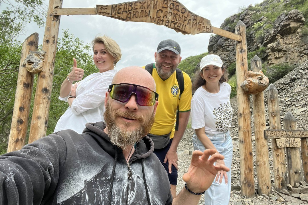 Группа из четырёх человек улыбается и позирует на фоне деревянной арки с надписью "Медвежий водопад". Люди выглядят счастливыми, одеты в светлую и спортивную одежду, очевидно, находятся в походе. За аркой видны склоны с зеленью и каменистая тропа. Атмосфера весёлая и дружелюбная, подчеркивает любовь к природе и приключениям.