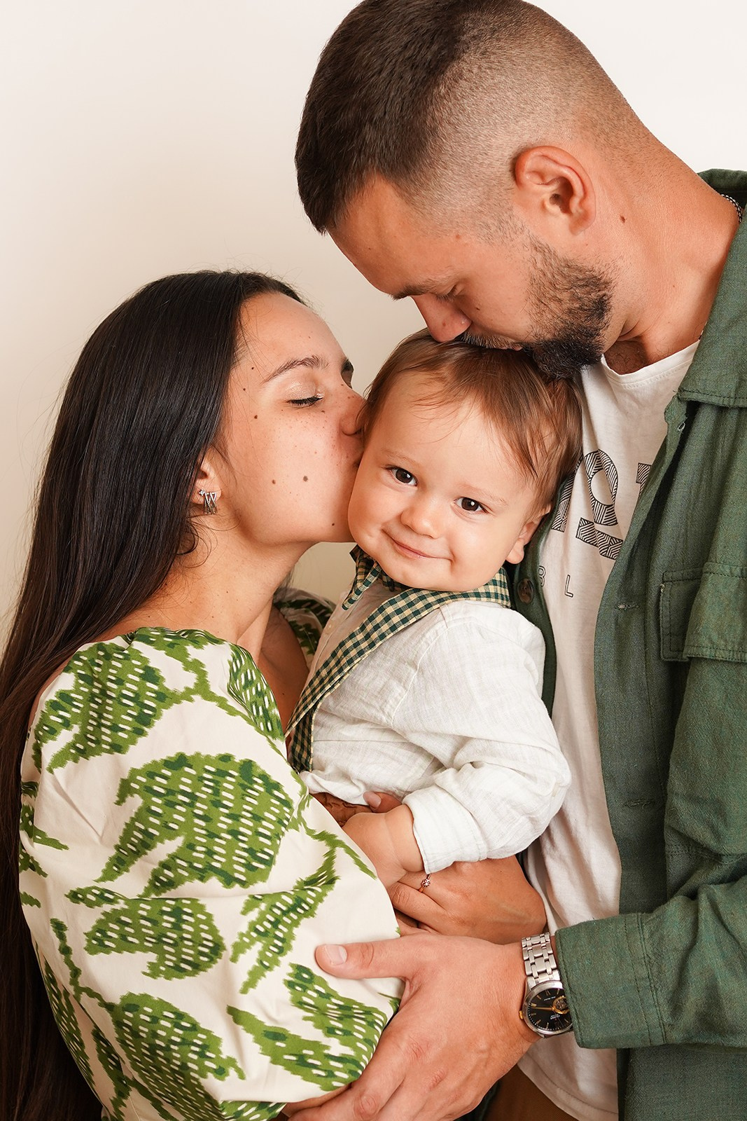 Семья приехала на фотосессию. На фото мама и папа целуют малыша. Он улыбается.