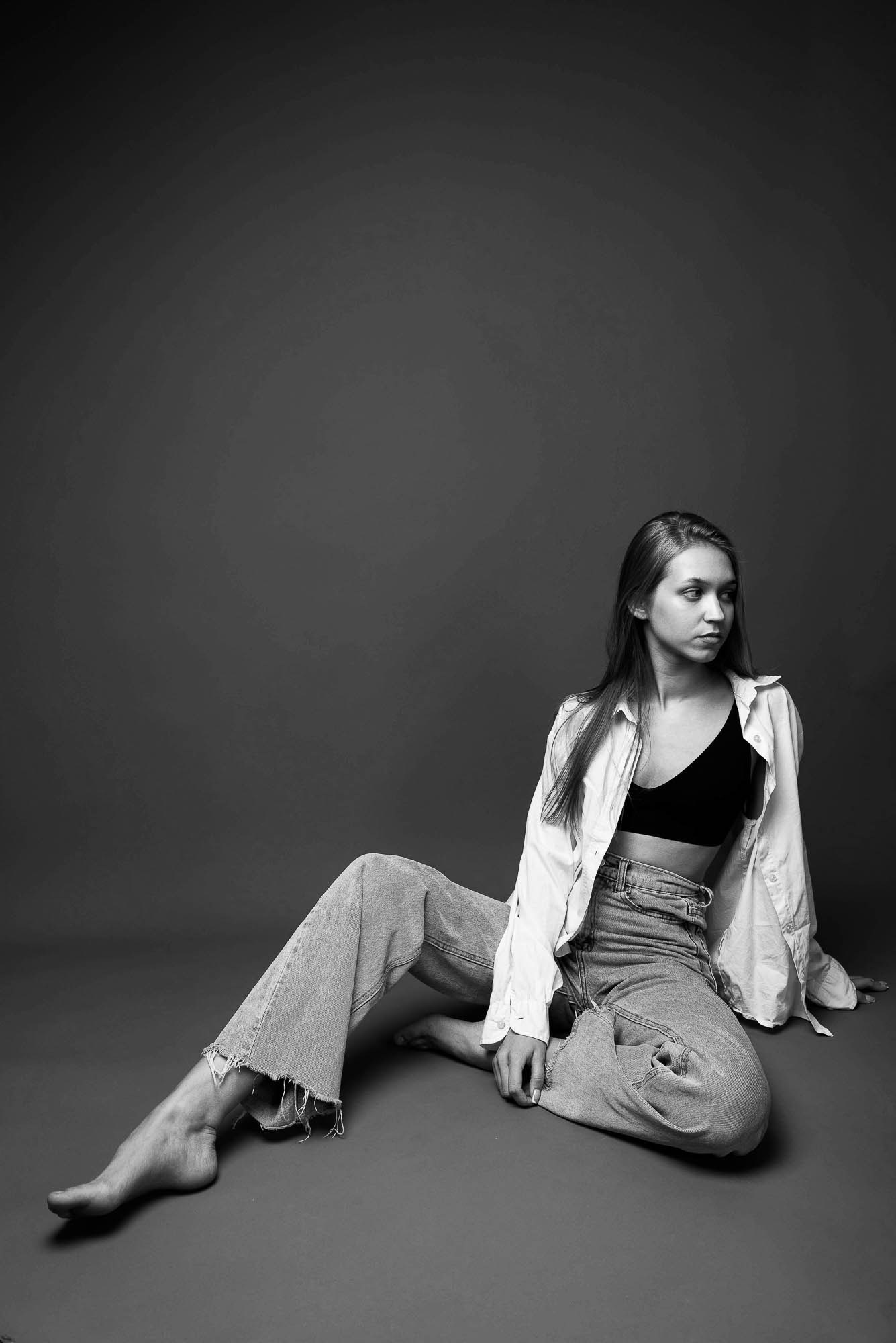Сидящая модель в чёрном топе и джинсах на чёрно-белом фоне, опирается на одну руку