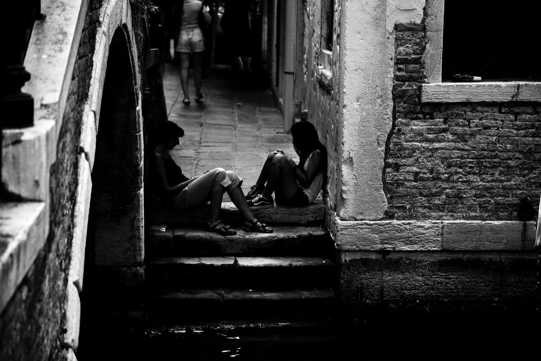 Черно-белое фото: двое людей сидят на каменных ступенях в узком венецианском переулке, арочный проход, старинная кирпичная кладка, люди проходят на заднем плане, уличная документальная фотография