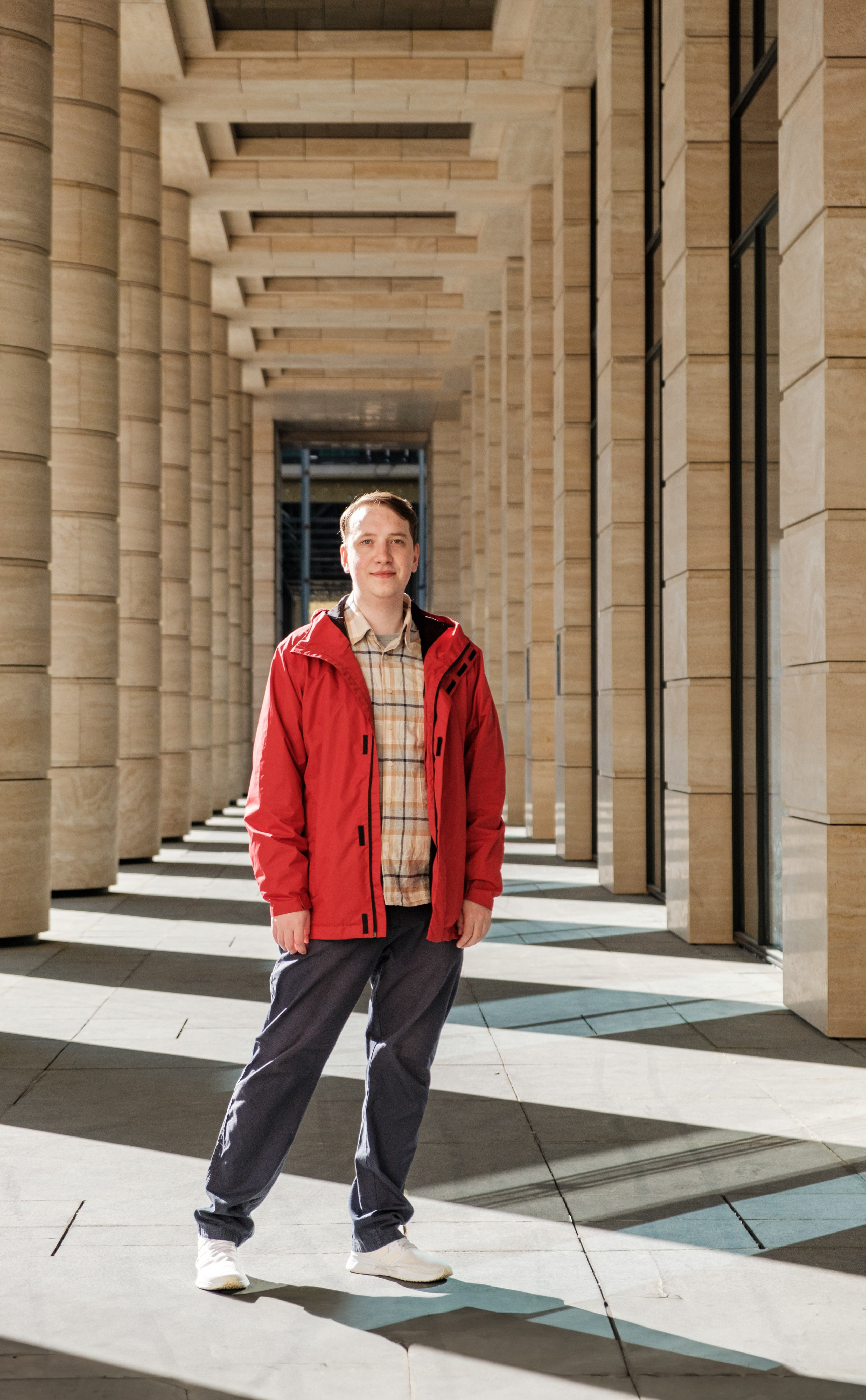 Молодой человек, стоящий внутри здания с колоннами. Он одет в красную куртку, тёмно-синие брюки и белые кроссовки.