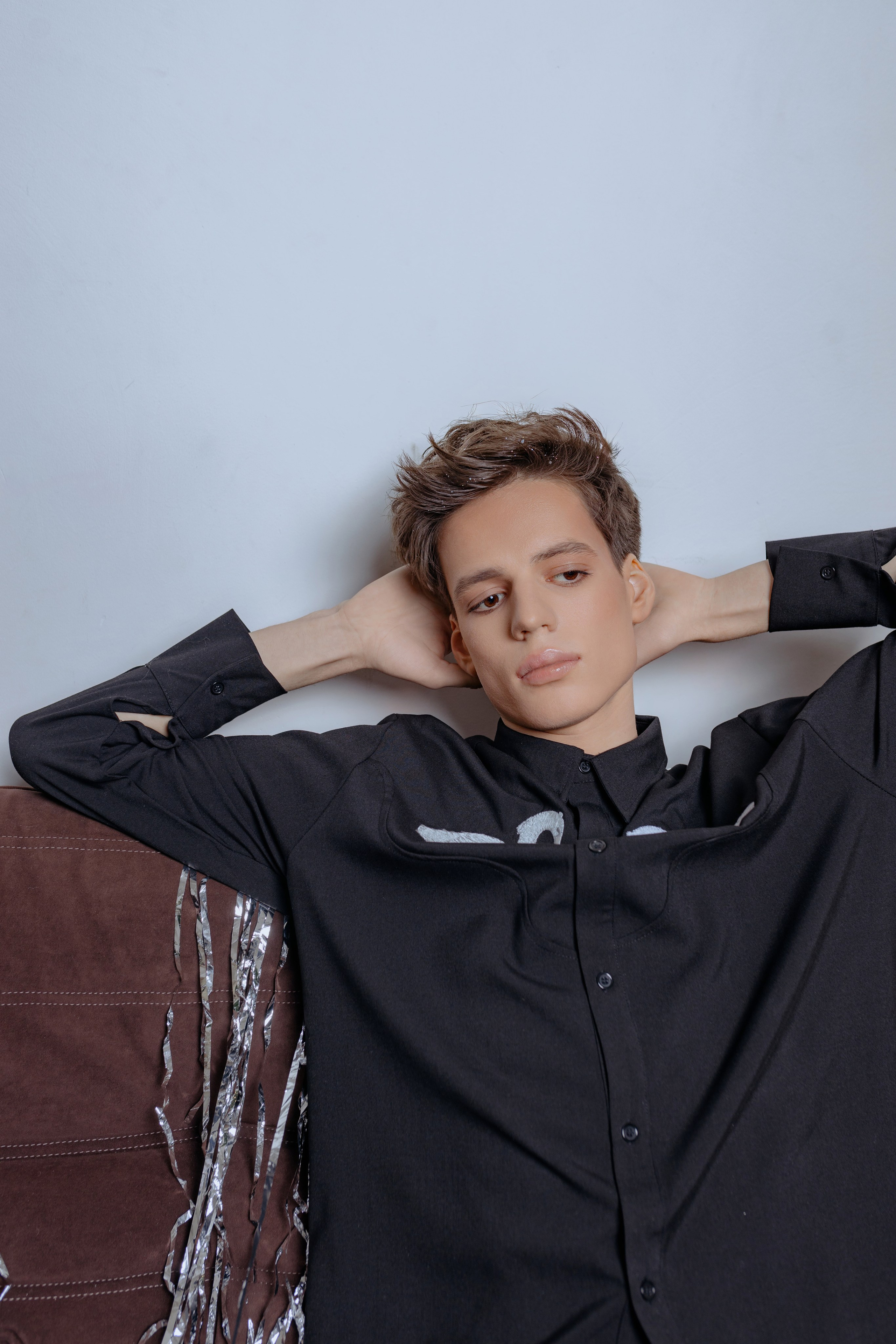 Парень в рубашке с лебедями и брюках с пайетками, блестки и стразы на лице. Fashion-портрет: стразы на лице, рубашка с принтом лебедей, взгляд в сторону от камеры, парень в сверкающих брюках и украшениях.