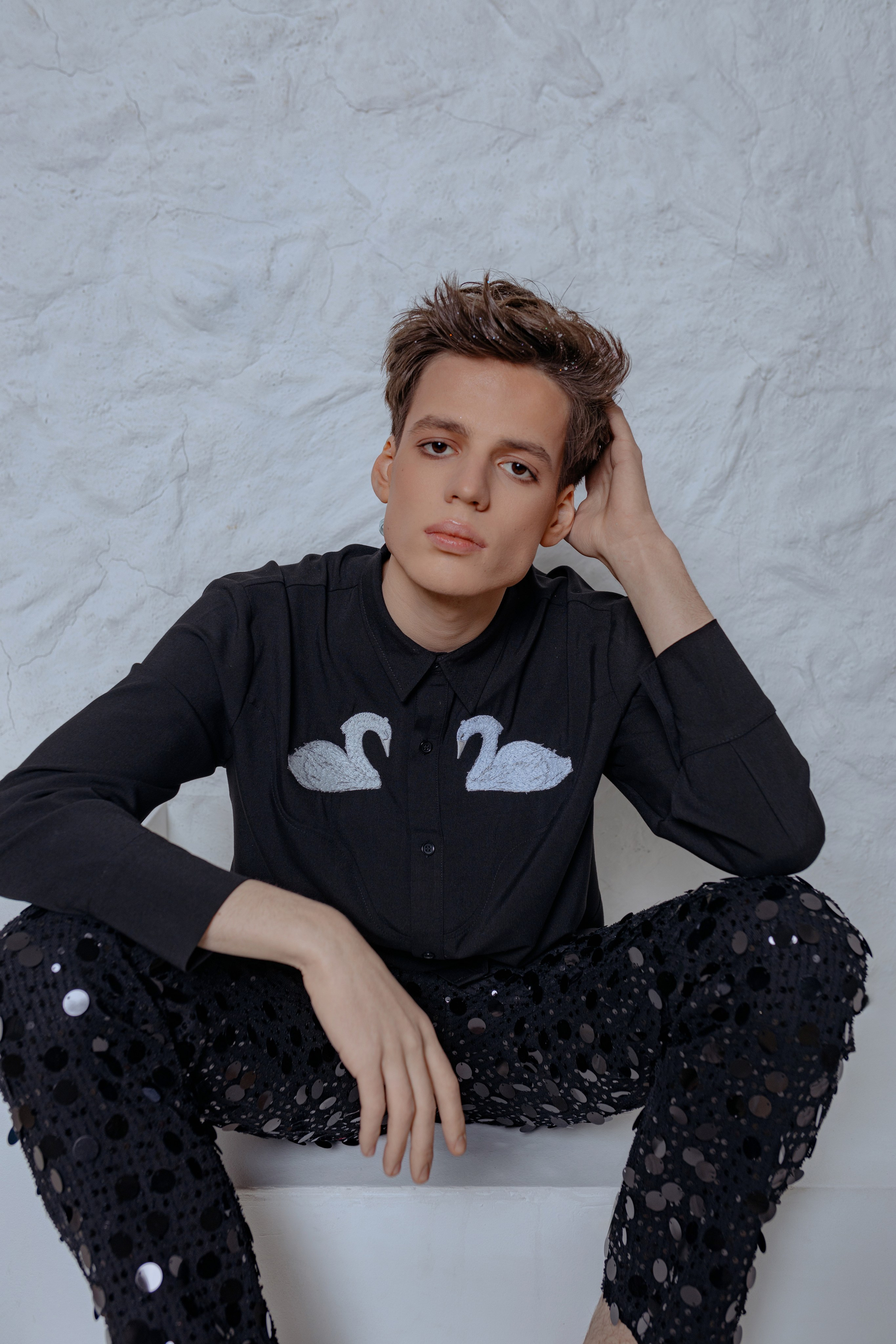 Парень в рубашке с лебедями и брюках с пайетками, блестки и стразы на лице. Fashion-портрет: стразы на лице, рубашка с принтом лебедей, взгляд в камеру, парень в сверкающих брюках и украшениях.