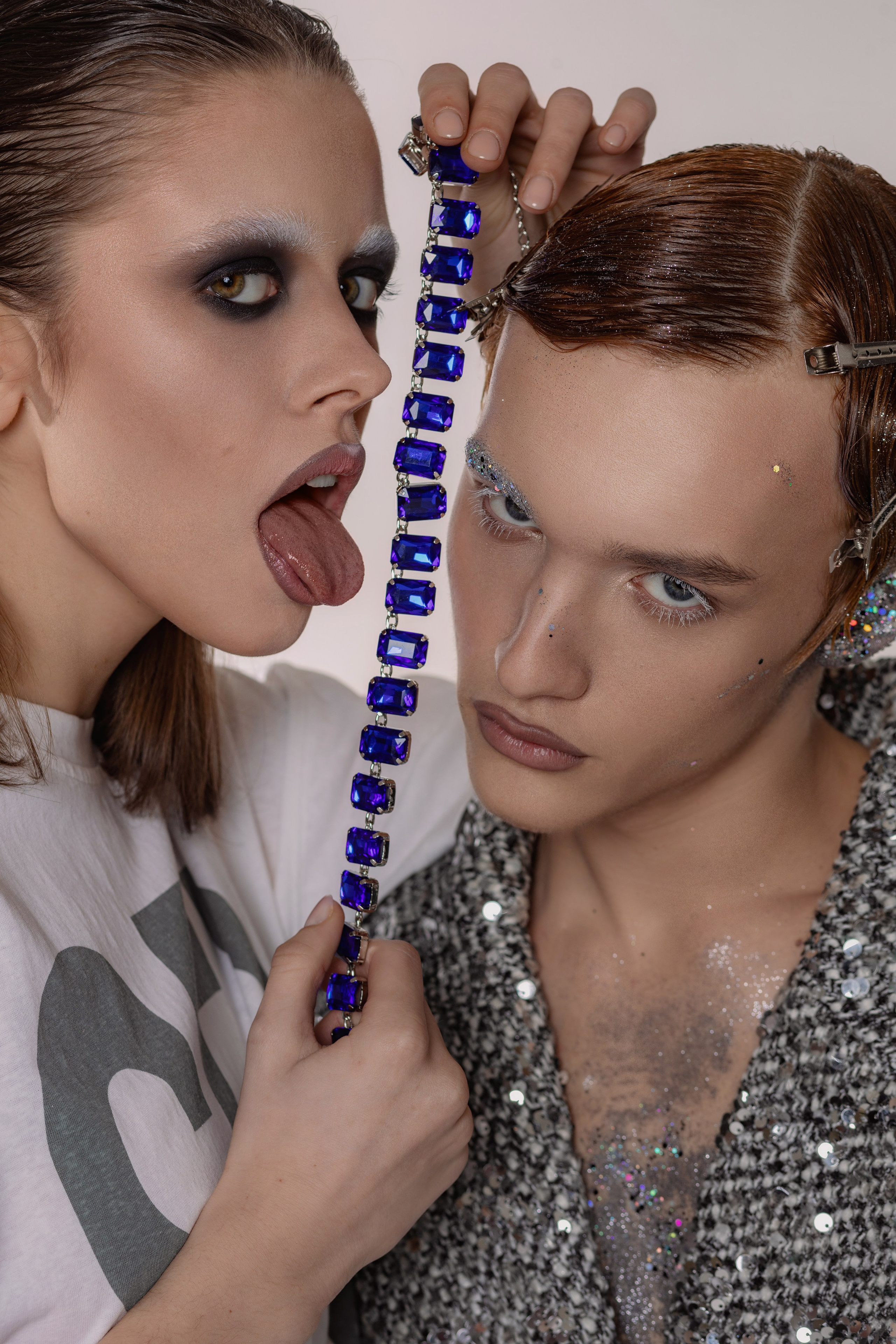 Девушка с колье из крупных камней и ярким макияжем, рядом парень с зализанными волосами. Портретная сцена: руки с колье, выразительный макияж, дуэт в сияющих образах. Модельная пара в королевской стилистике: акценты на украшениях и макияже. 