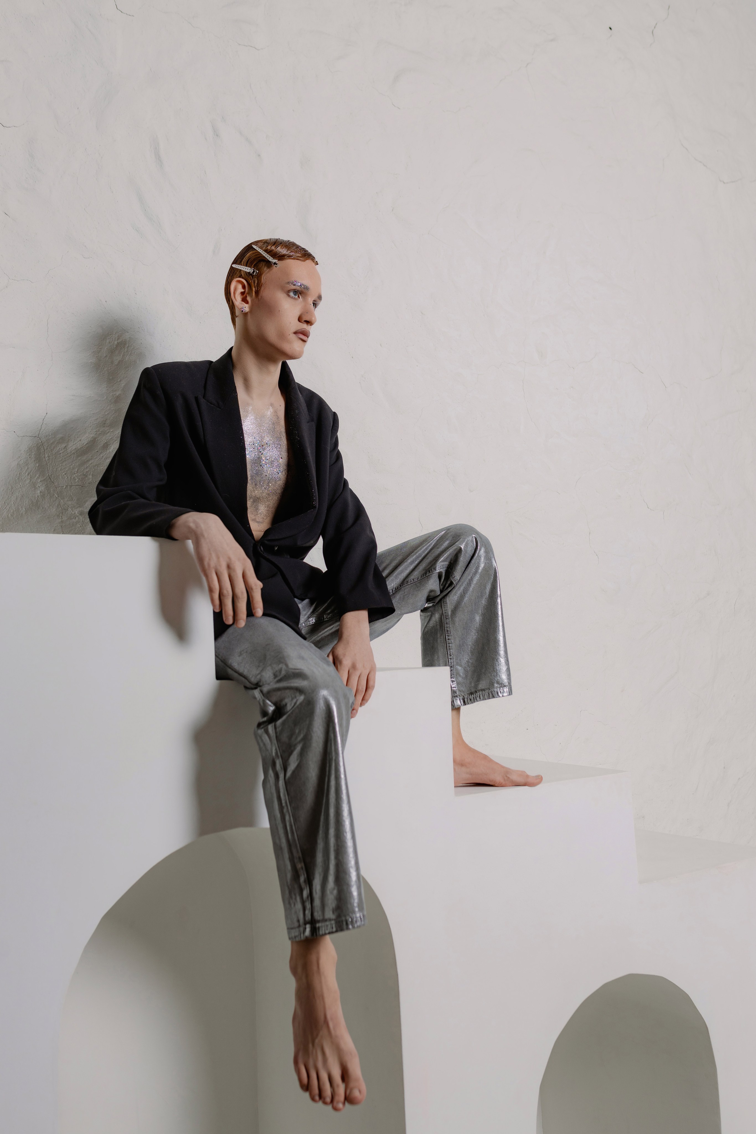 Парень с зализанными волосами в пиджаке на голое тело и брюках металлик. Образ в стиле новой роскоши: сияющая ткань, обнажённая грудь, уверенный взгляд. Fashion-съёмка: мужская модель в наряде металлик на фоне нейтрального фона. 