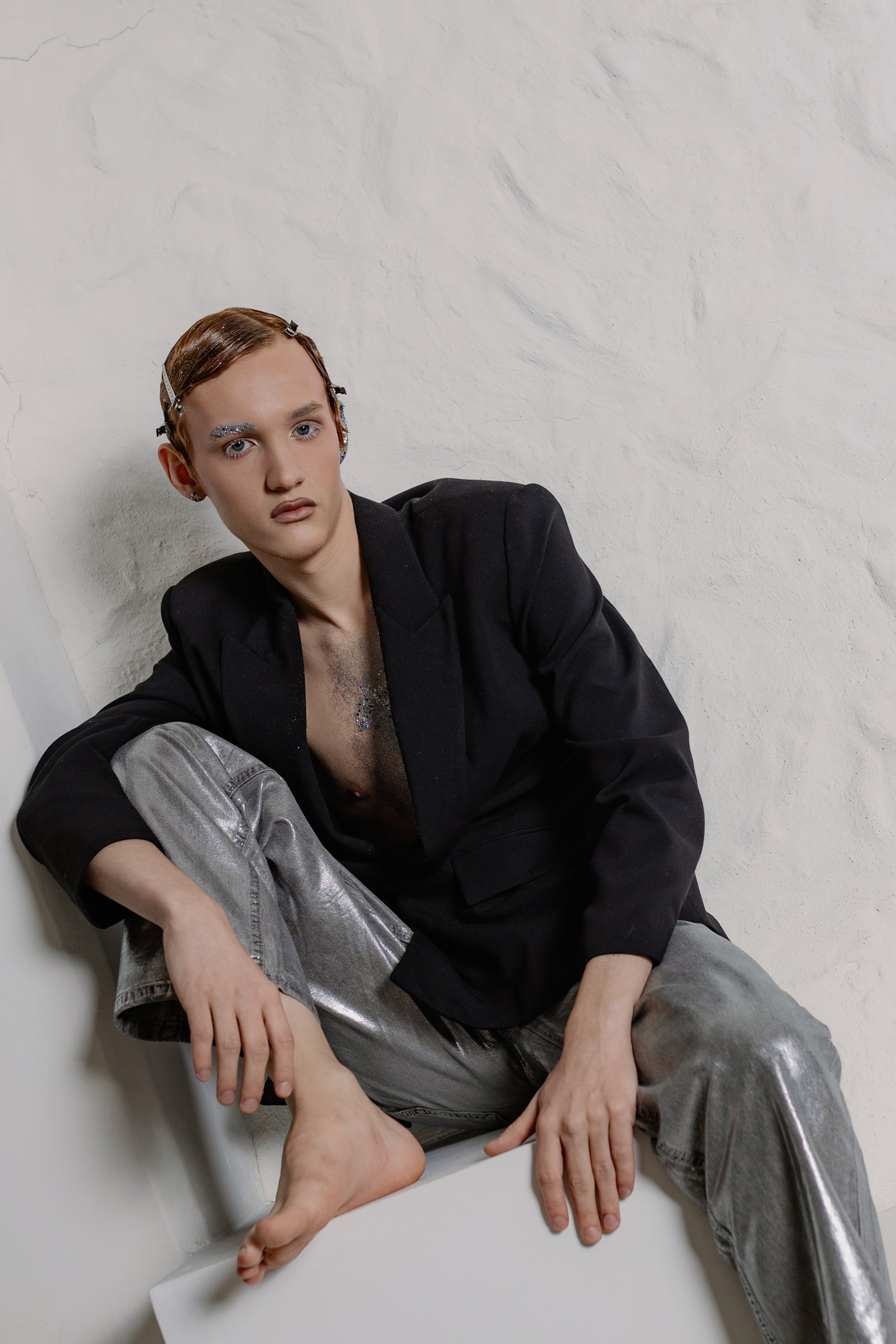 Парень с зализанными волосами в пиджаке на голое тело и брюках металлик. Образ в стиле новой роскоши: сияющая ткань, обнажённая грудь, уверенный взгляд. Fashion-съёмка: мужская модель в наряде металлик на фоне нейтрального фона. 