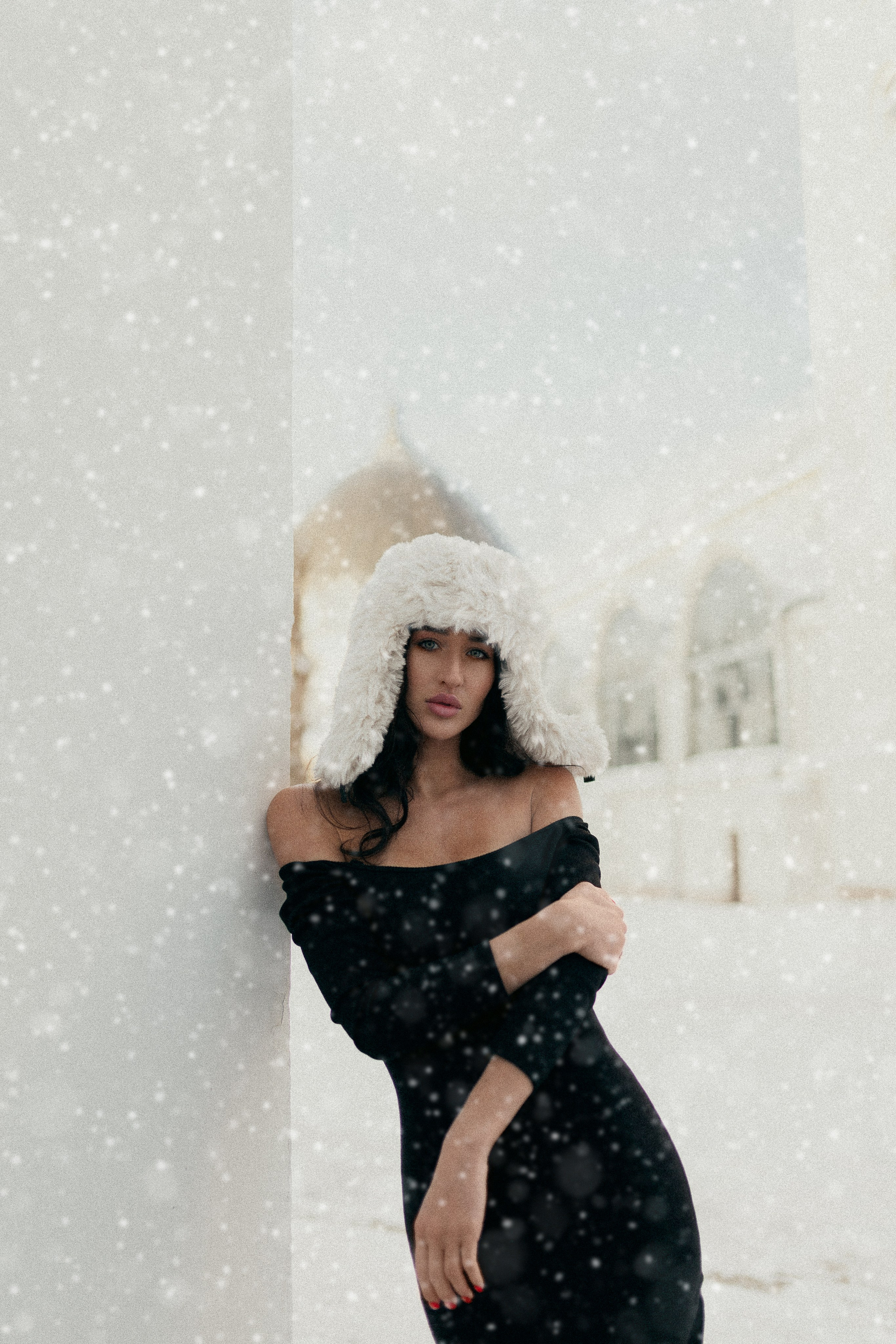 Девушка в черном платье и в белой меховой шапке на фоне заснеженного города