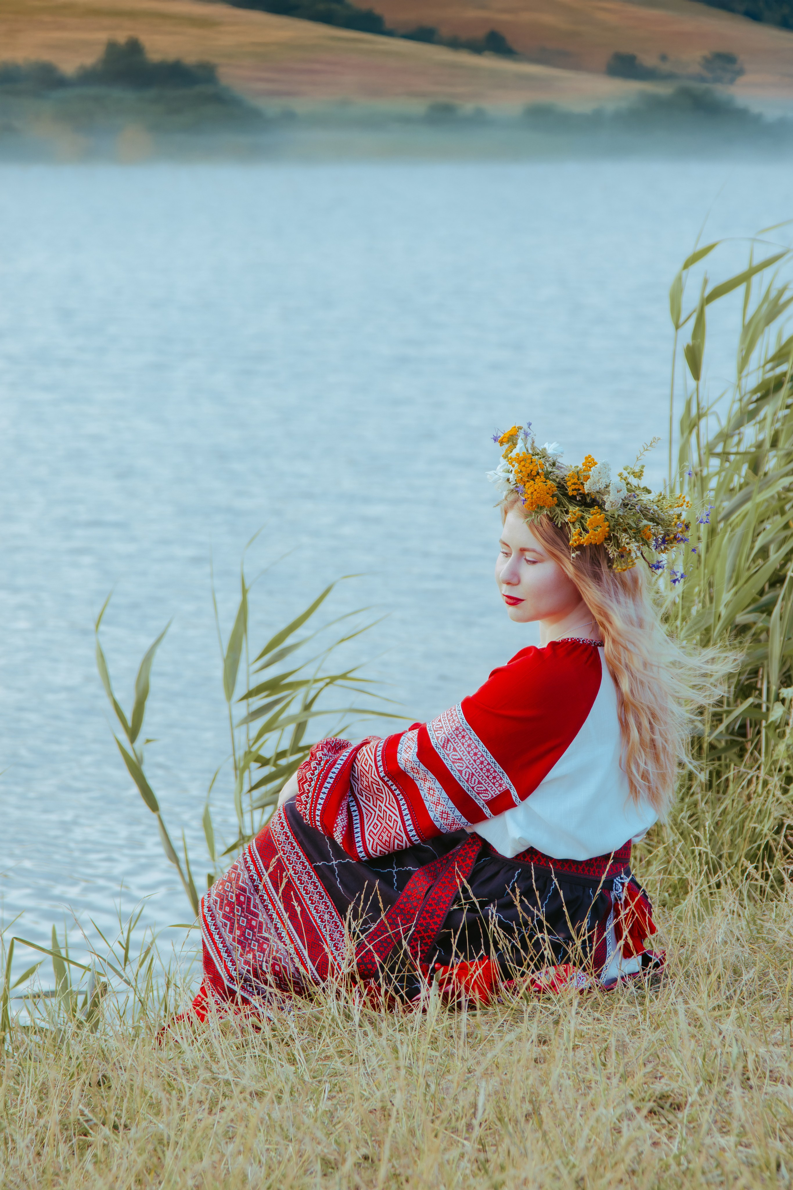 Славянская девушка сидит на берегу реки с венком на голове и смотрит в дль