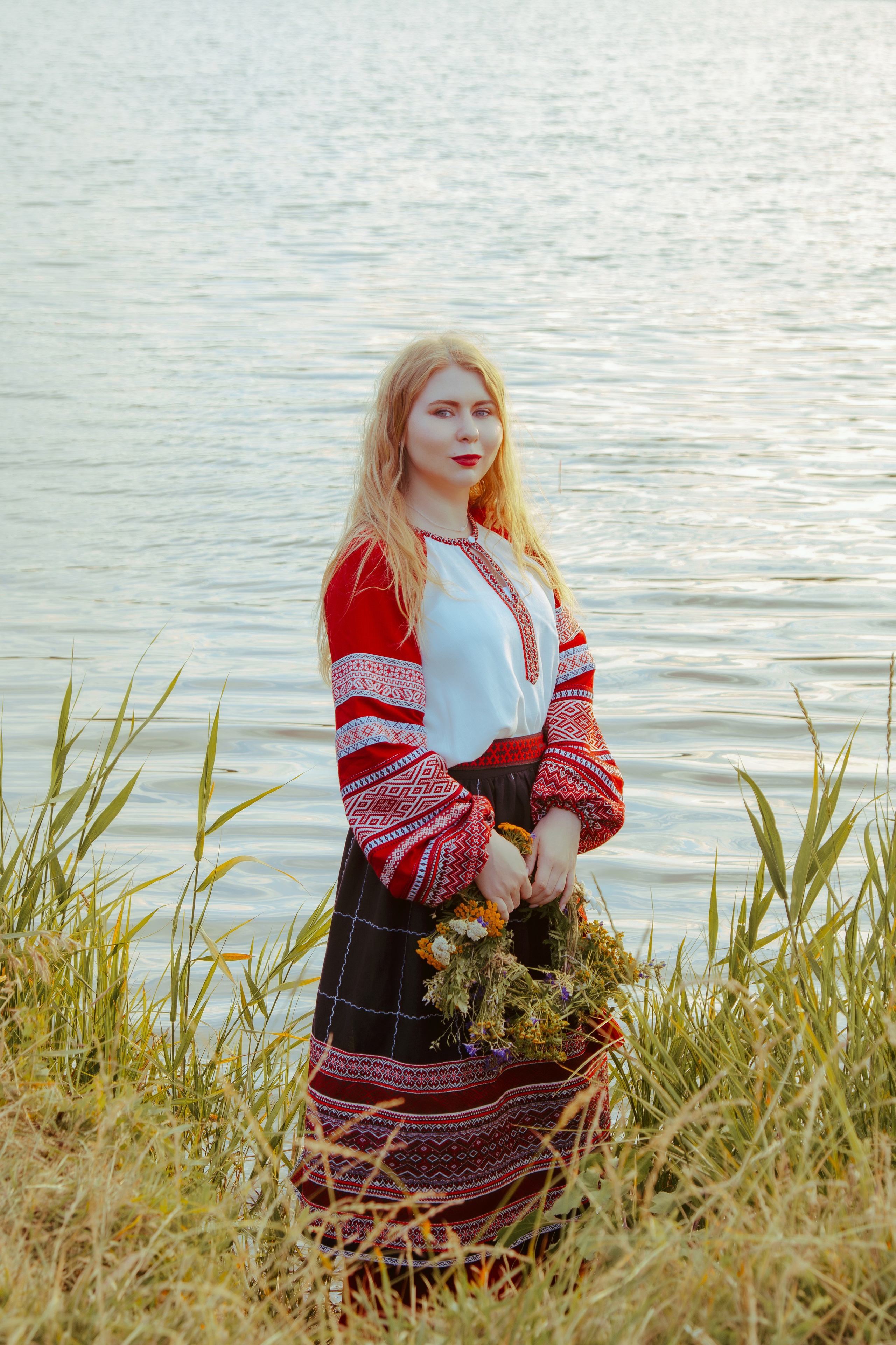 Девушка стоит на фоне реки в лесу, в руках держа венок из цветов