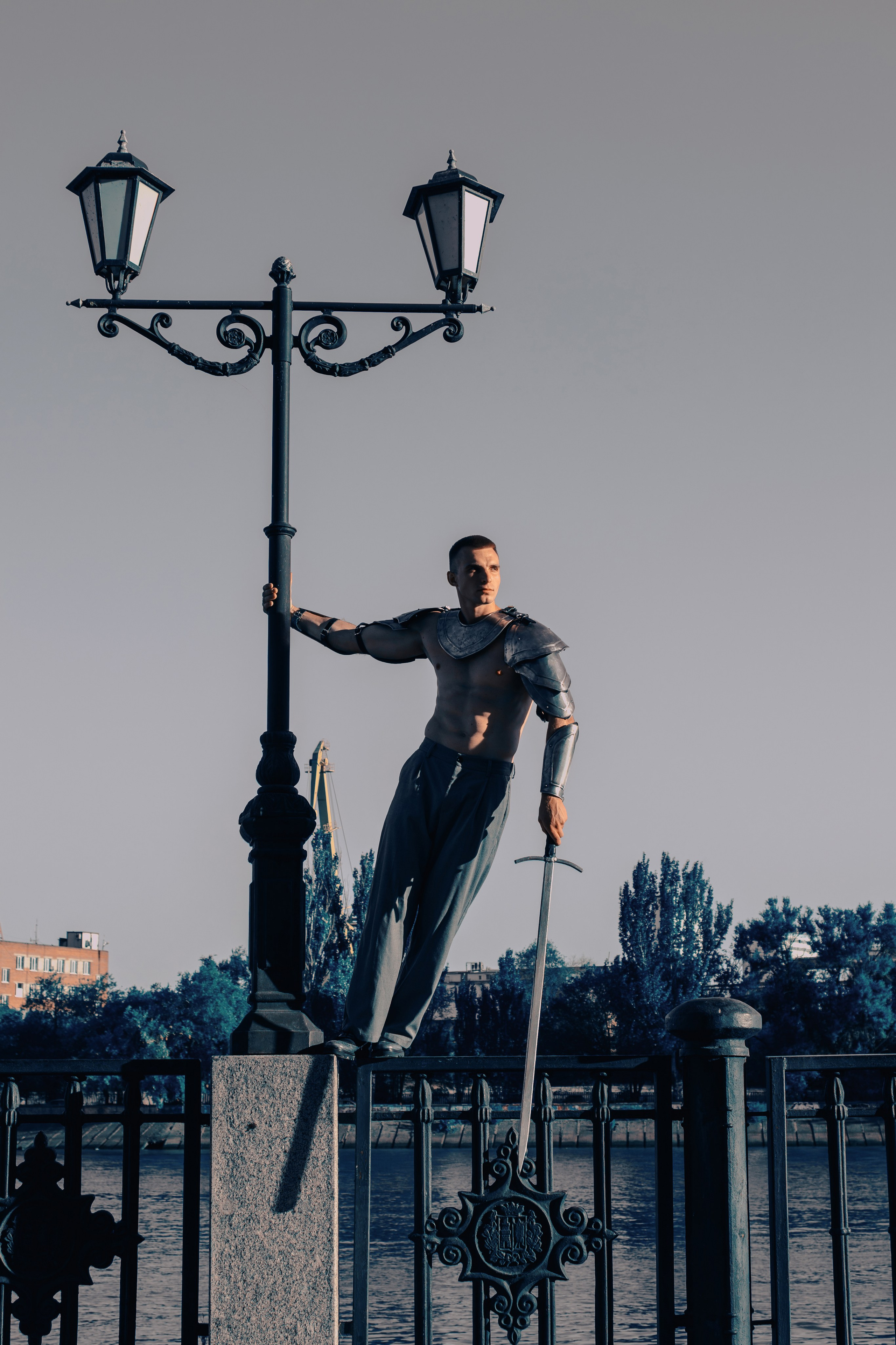 В полный рост в доспехах, брюках и туфлях стоит на заборе мужчина с мечом в руках