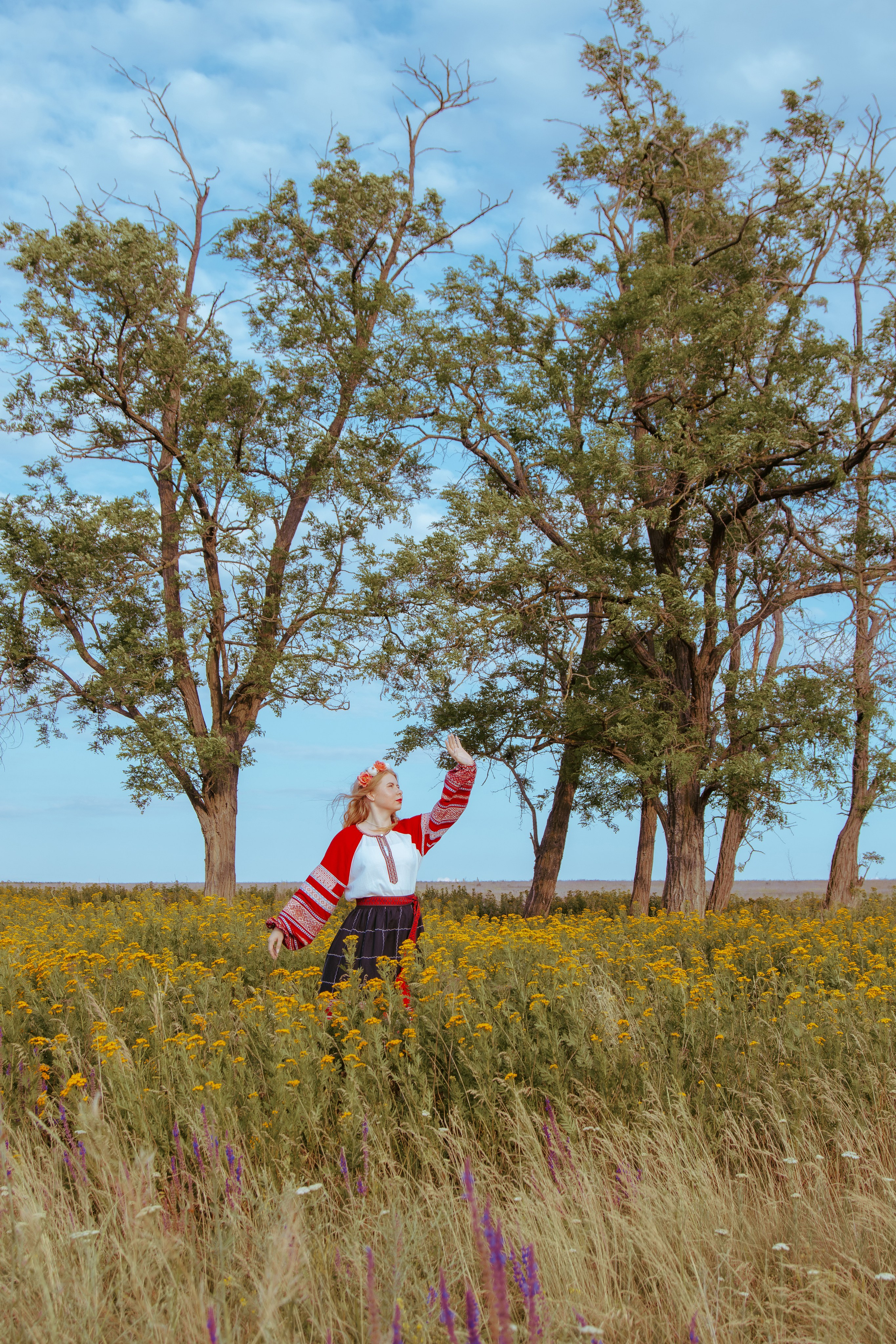 Девушка в красном славянском платье стоит на фоне деревьев в поле