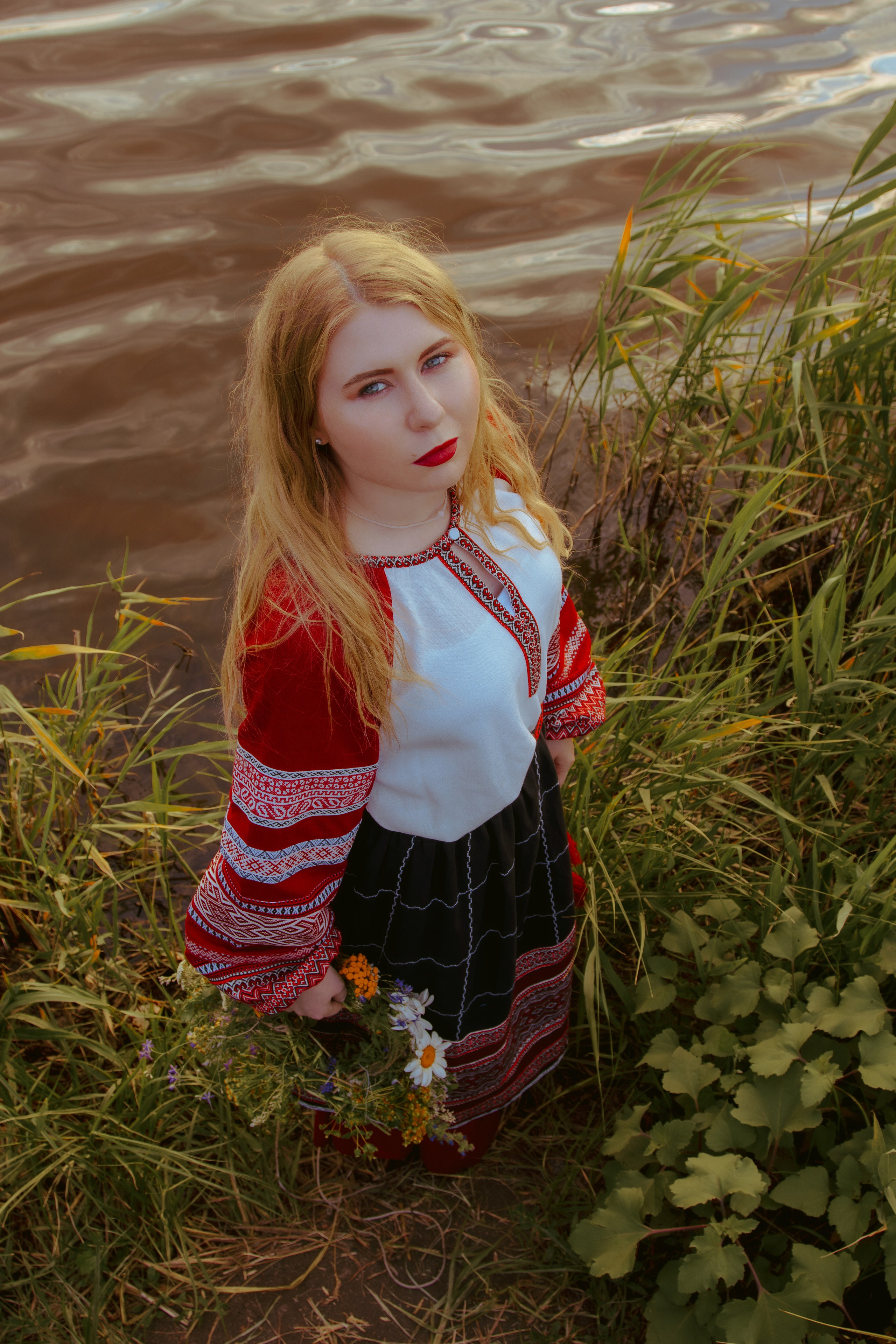 Девушка в славянской одежде стоит на фоне реки с грустным лицом 