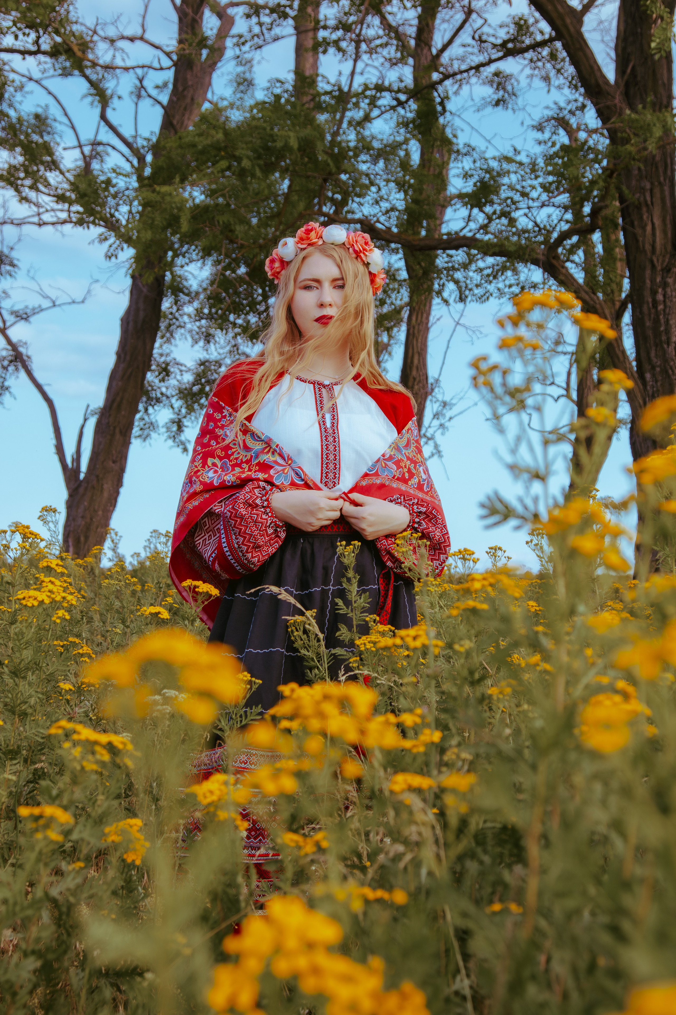 На фоне деревьев стоит девушка в красном славянском платье держа свои руки на поясе