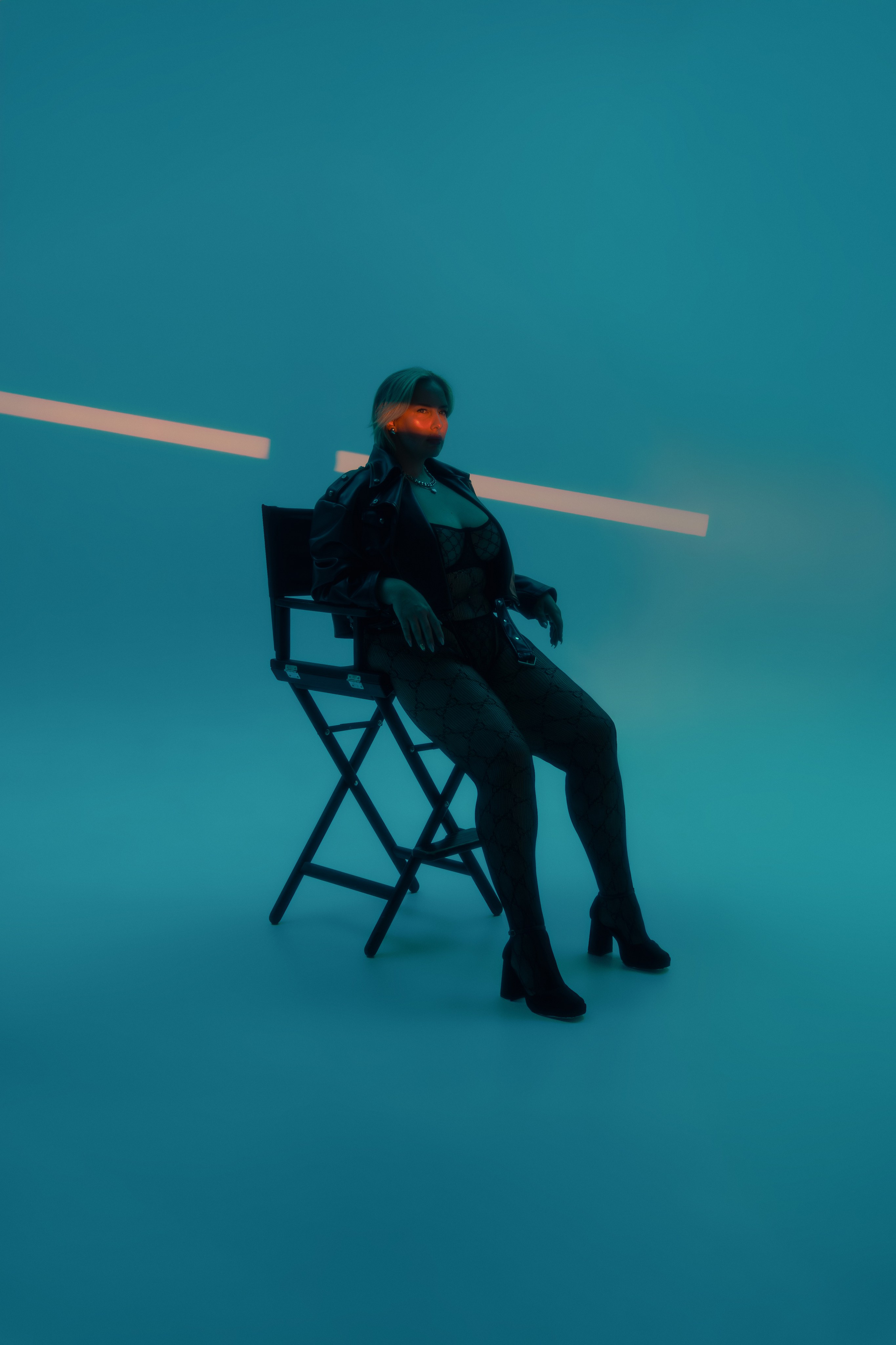 Модель плюс сайз на синем фоне с оранжевой полоской света на лице позирует на стуле