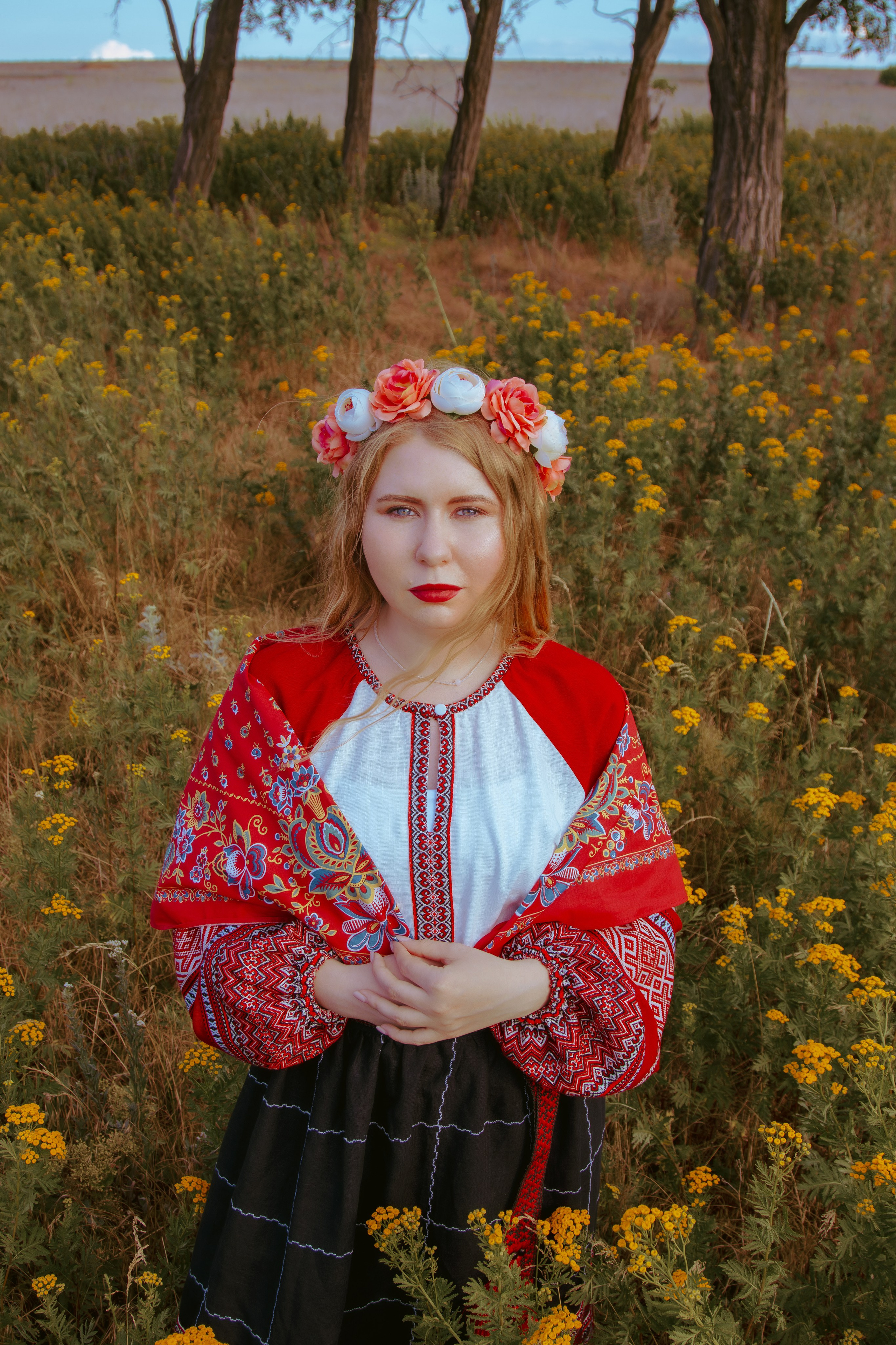 девушка в красном платье славянского стиля стоит в поле ромашек и смотрит вверх 