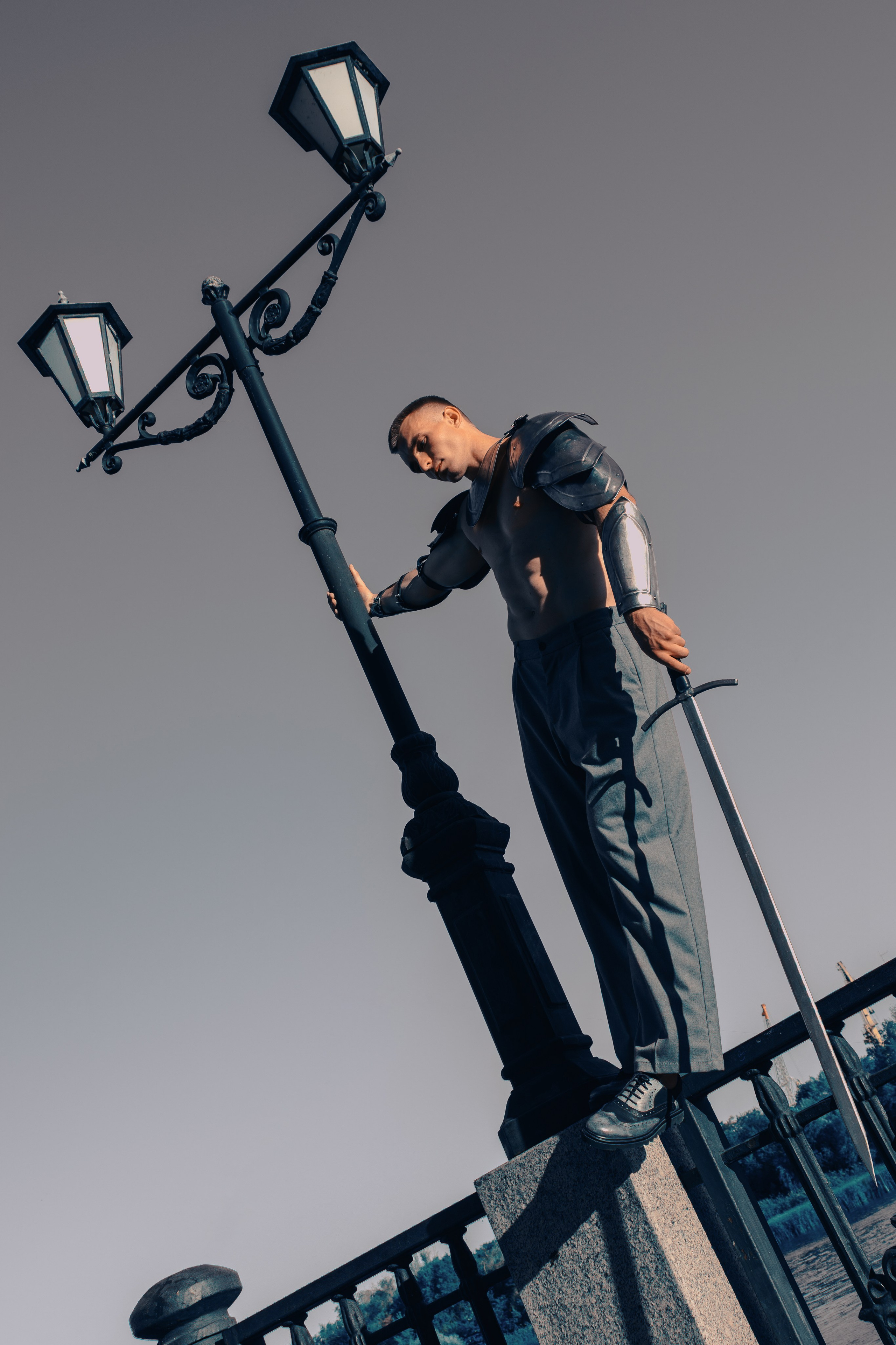 В полный рост в доспехах, брюках и туфлях стоит на заборе мужчина с мечом в руках