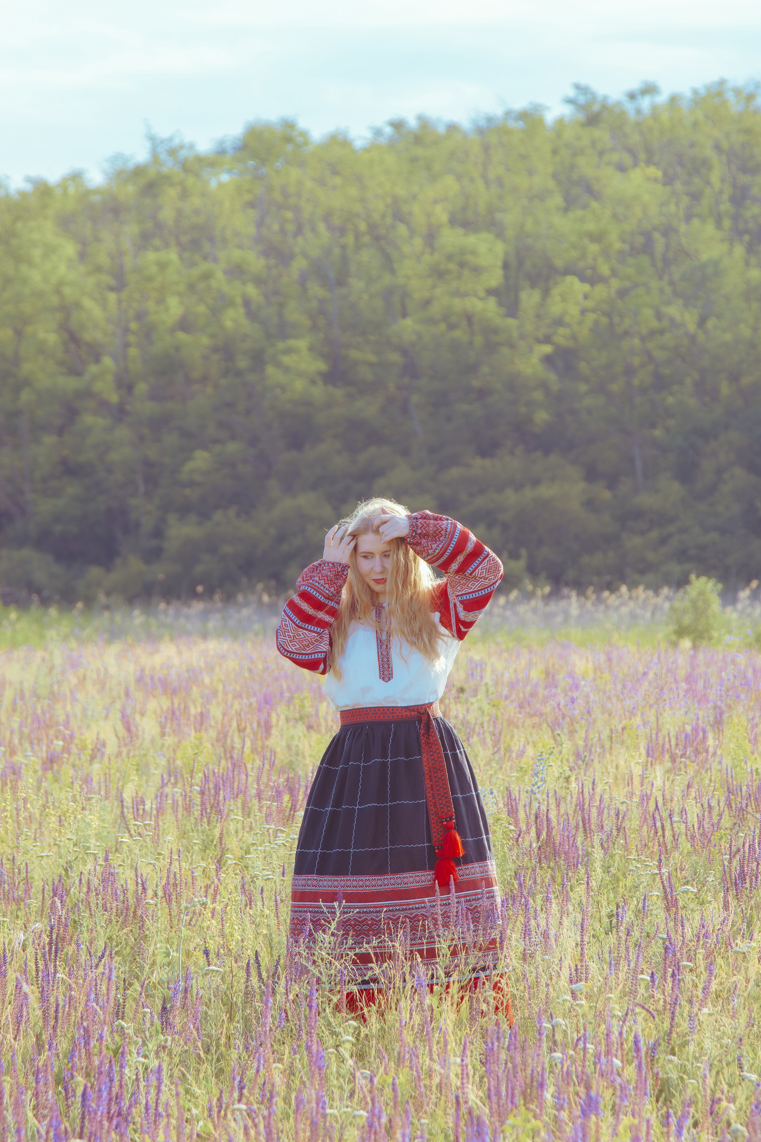 Славянская девушка стоит в поле на фоне леса и солнечными лучами 