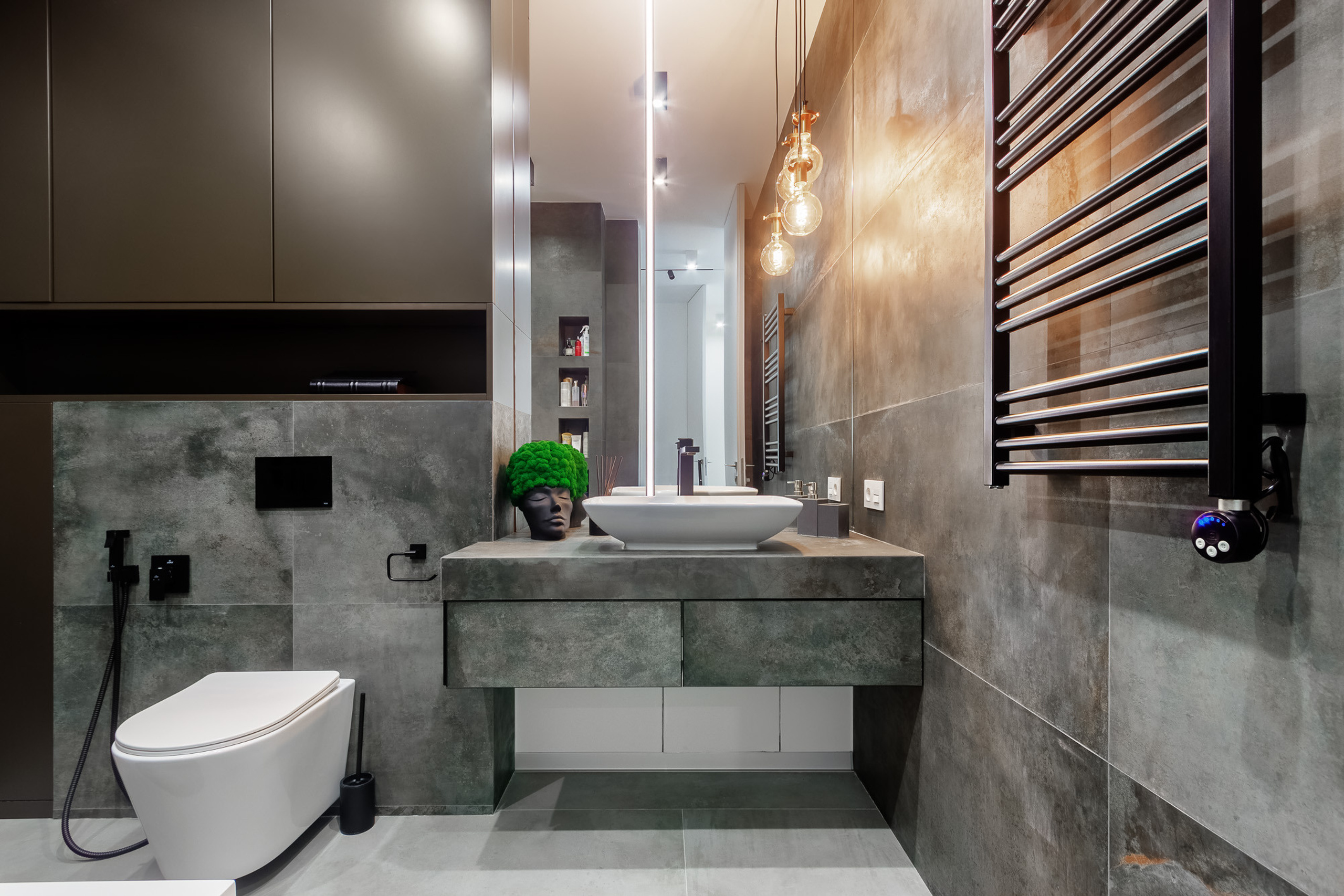 Умывальник, полотенцесушитель и унитаз в большой тёмно-серой ванной комнате с тёплым жёлтым освещением