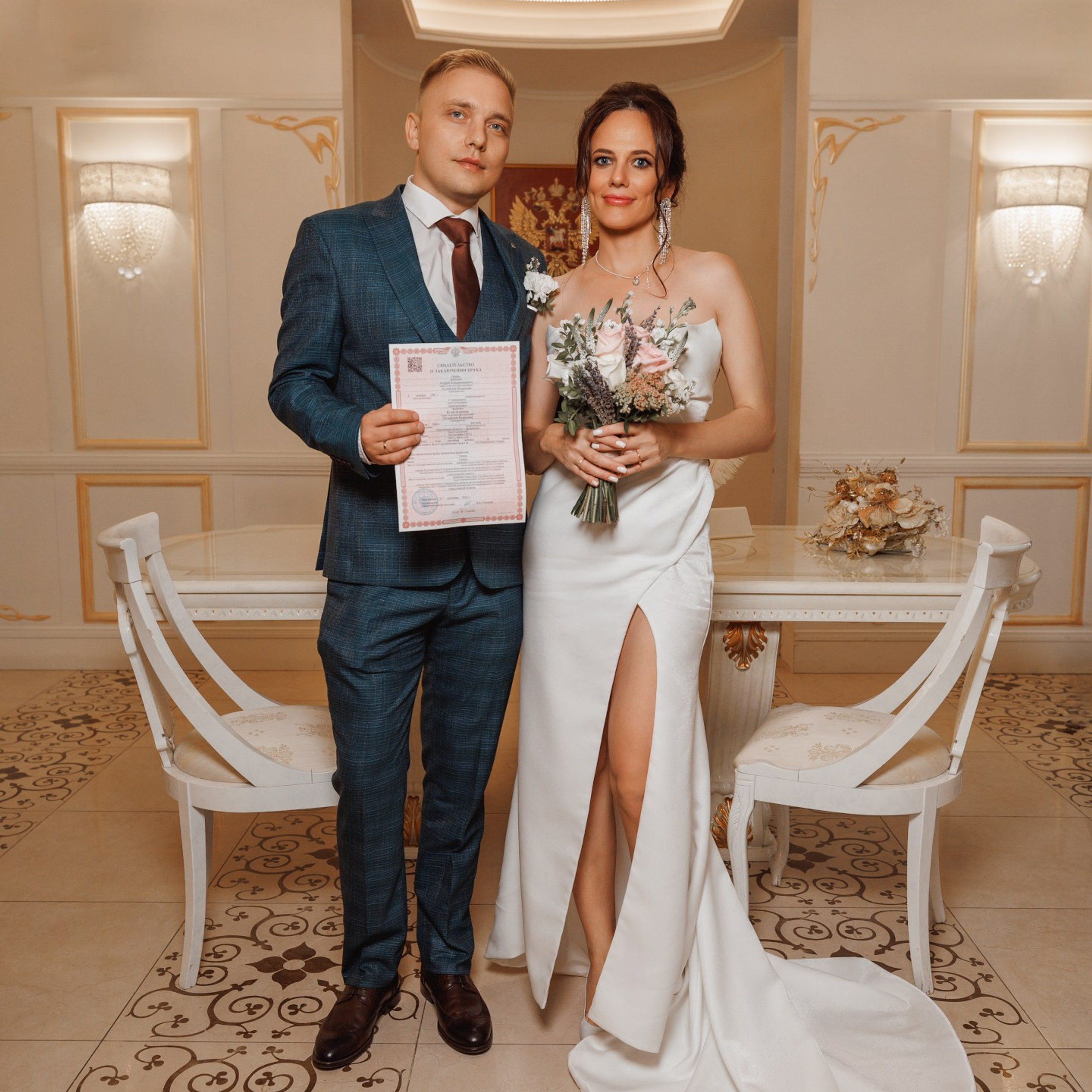 Молодожёны позируют в зале бракосочетаний ЗАГСа со свидетельством о браке в руках. На фото запечатлён торжественный момент после официальной церемонии, где пара радостно демонстрирует документ, символизирующий начало их семейной жизни.