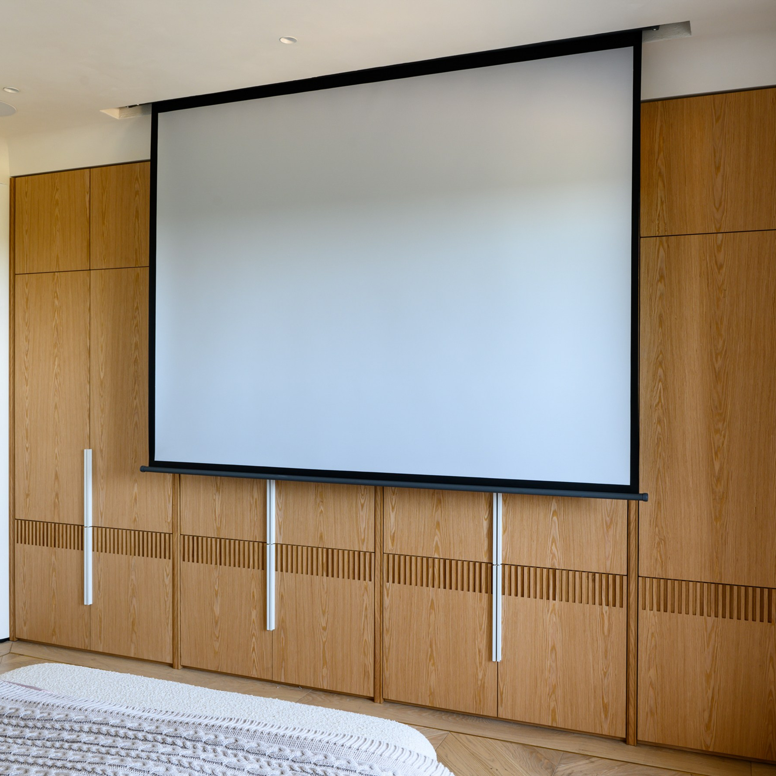 Длинную сторону комнаты займёт многофункциональная система хранения. Перед ней, в нише, разместится моторизованный потолочный экран для проектора. Кроме того, мы постараемся использовать моторизованные карнизы для портьер с подкладом из ткани блэк-аут.