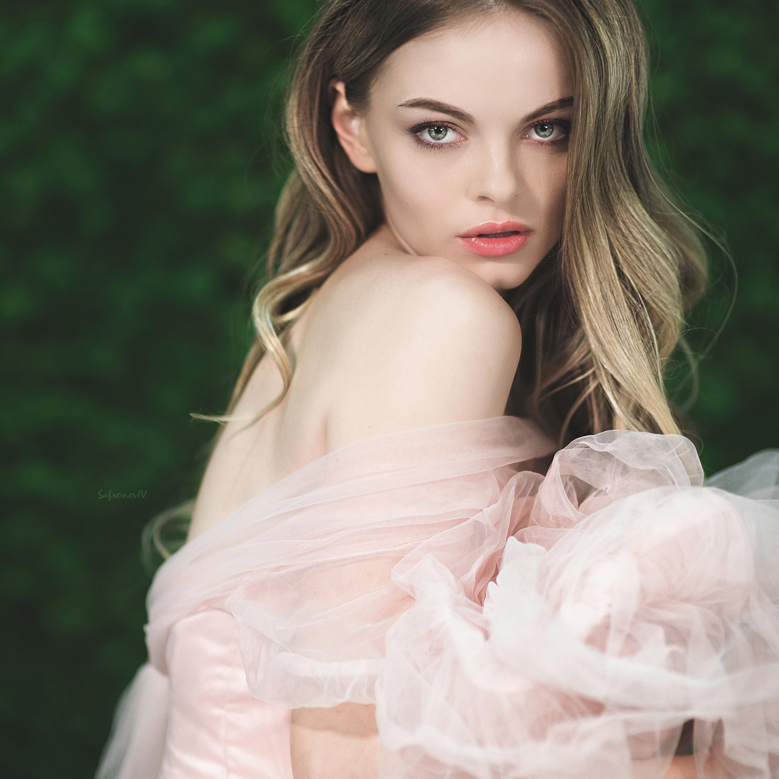 молодая женщина в нежно-розовом платье с воздушными, полупрозрачными рукавами. Её длинные светлые волосы волнами ниспадают на плечи, подчёркивая утончённость образа. Взгляд модели направлен через плечо — он глубокий, немного загадочный, будто приглашает зрителя в мир её мыслей. Макияж выполнен в естественных тонах, акцентируя внимание на выразительных глазах и нежных губах. Фон — размытый, насыщенного зелёного цвета, создаёт контраст с нежностью наряда и кожи модели, подчёркивая её хрупкость и элегантность. Композиция кадра подчёркивает грацию и женственность героини.Заголовок:«Нежность в каждом изгибе: портрет утончённой красоты»Ключевые слова: портретная фотосессия, элегантность, нежность, эстетика, розовое платье, женственность, студийная съёмка, портретный фотограф в Москве, фотограф Сафронов Иван.