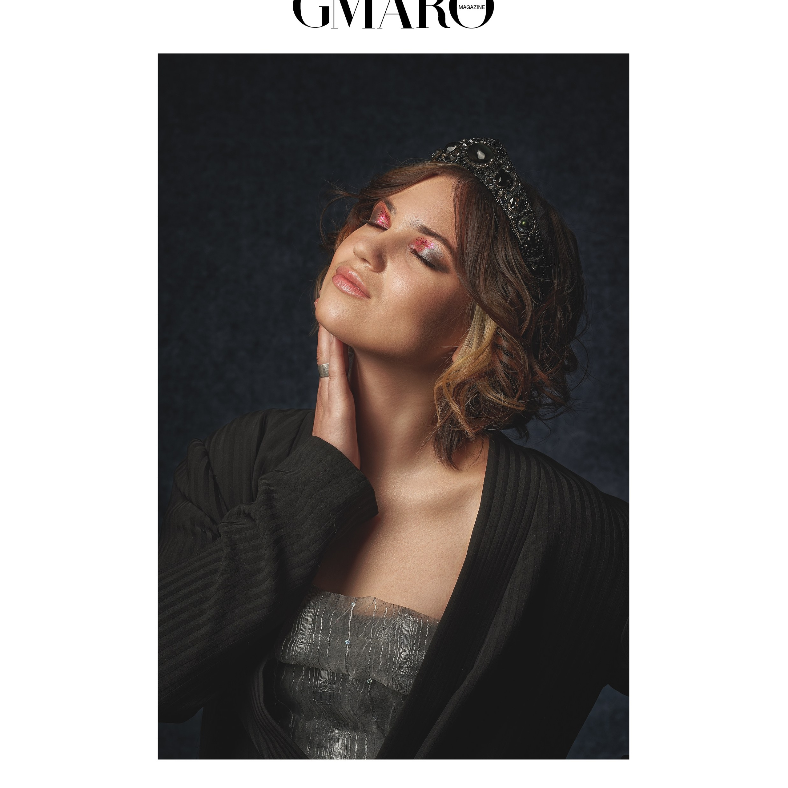 На фото — обложка издания GMARO Magazine. В центре кадра — молодая женщина в расслабленной, но выразительной позе: голова слегка запрокинута, глаза закрыты, пальцы мягко касаются шеи. Образ сочетает сдержанную элегантность и артистичную чувственность. На девушке — фактурный серебристый топ без бретелей, поверх накинут объёмный чёрный кардиган в рубчик. Причёска — мягкие локоны до плеч, а завершает образ тёмная декоративная диадема с камнями: она добавляет нотку «королевской» утончённости. Макияж выполнен в пастельно‑розовой гамме с лёгким шиммером, подчёркивая хрупкость и мечтательность образа. Глубокий тёмно‑синий фон и мягкий направленный свет создают кинематографичный контраст, выделяя силуэт и текстуру тканей.Варианты заголовков на русском:«Тихая роскошь: портрет в полутени»«Миг безмятежности: элегантность в деталях»«Королевская небрежность: искусство расслабленного портрета»«Между сном и явью: чувственный портрет для обложки»«Элегантность в полумраке: игра света и фактур»Ключевые слова по фото:обложка журнала, студийный портрет, элегантная фотосессия, диадема, фактурные ткани, мягкий свет, портрет в полутени, художественная съёмка, фотосессия в Москве, портретный фотограф в Москве, фотограф Сафронов Иван.