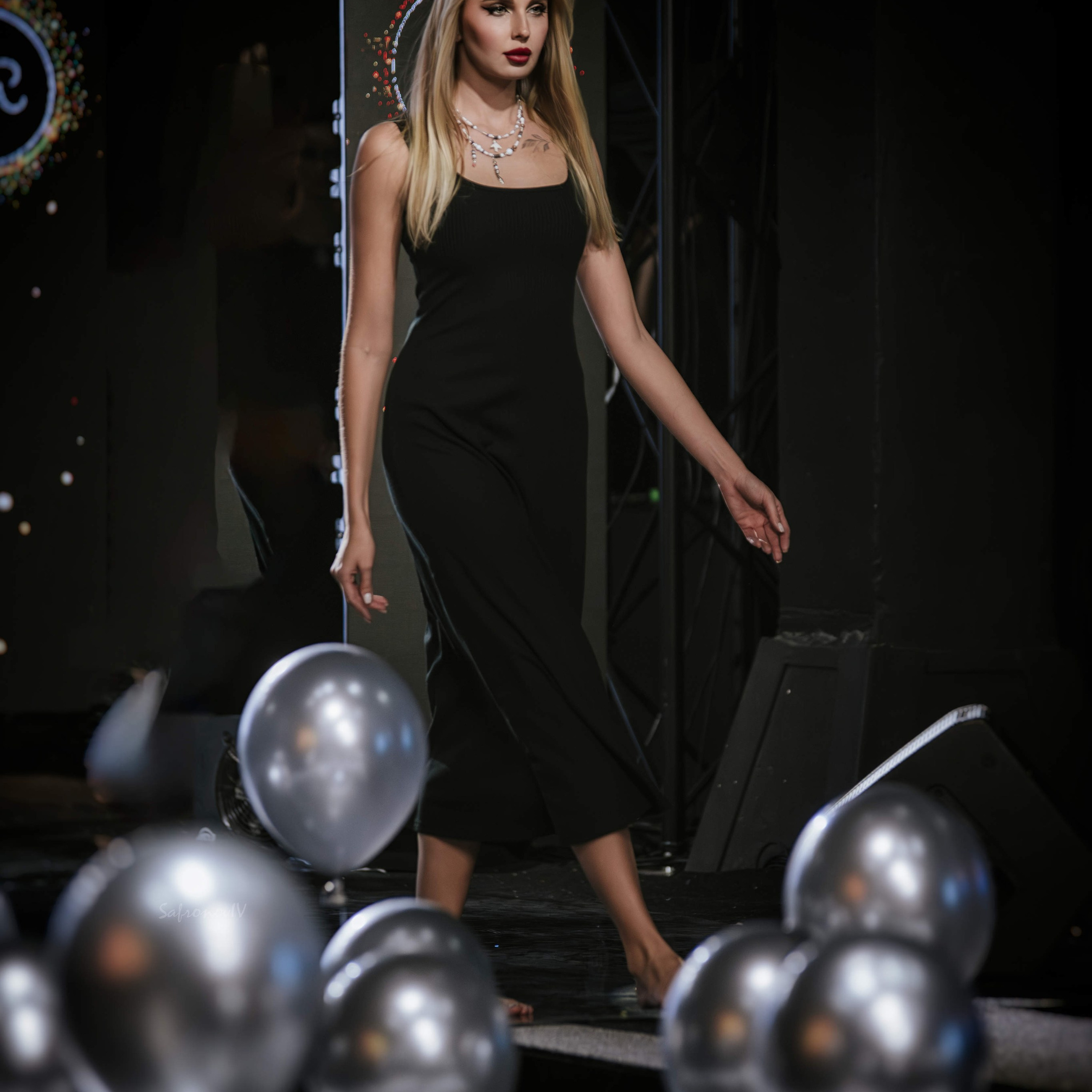 молодая женщина в элегантном чёрном платье, стоящая на сцене. Её светлые волосы аккуратно уложены, а макияж подчёркивает выразительность взгляда. Модель выглядит уверенно и грациозно: поза расслабленная, но в то же время демонстрирует утончённость и стиль. На шее у неё изящное украшение — цепочка с подвеской.На переднем плане — группа серебристых шаров, которые создают интересный визуальный акцент и добавляют снимку праздничности. Фон затемнён, но можно различить элементы сценического декора: световые эффекты и конструкции, создающие атмосферу шоу или модного показа. Освещение сфокусировано на модели, подчёркивая её силуэт и детали образа.Заголовок«Элегантность на сцене: игра света и стиля»Ключевые словаэлегантность, сцена, чёрное платье, портретная съёмка, фотосессия, серебристые шары, сценический образ, стиль, портретный фотограф в Москве, фотограф Сафронов Иван.