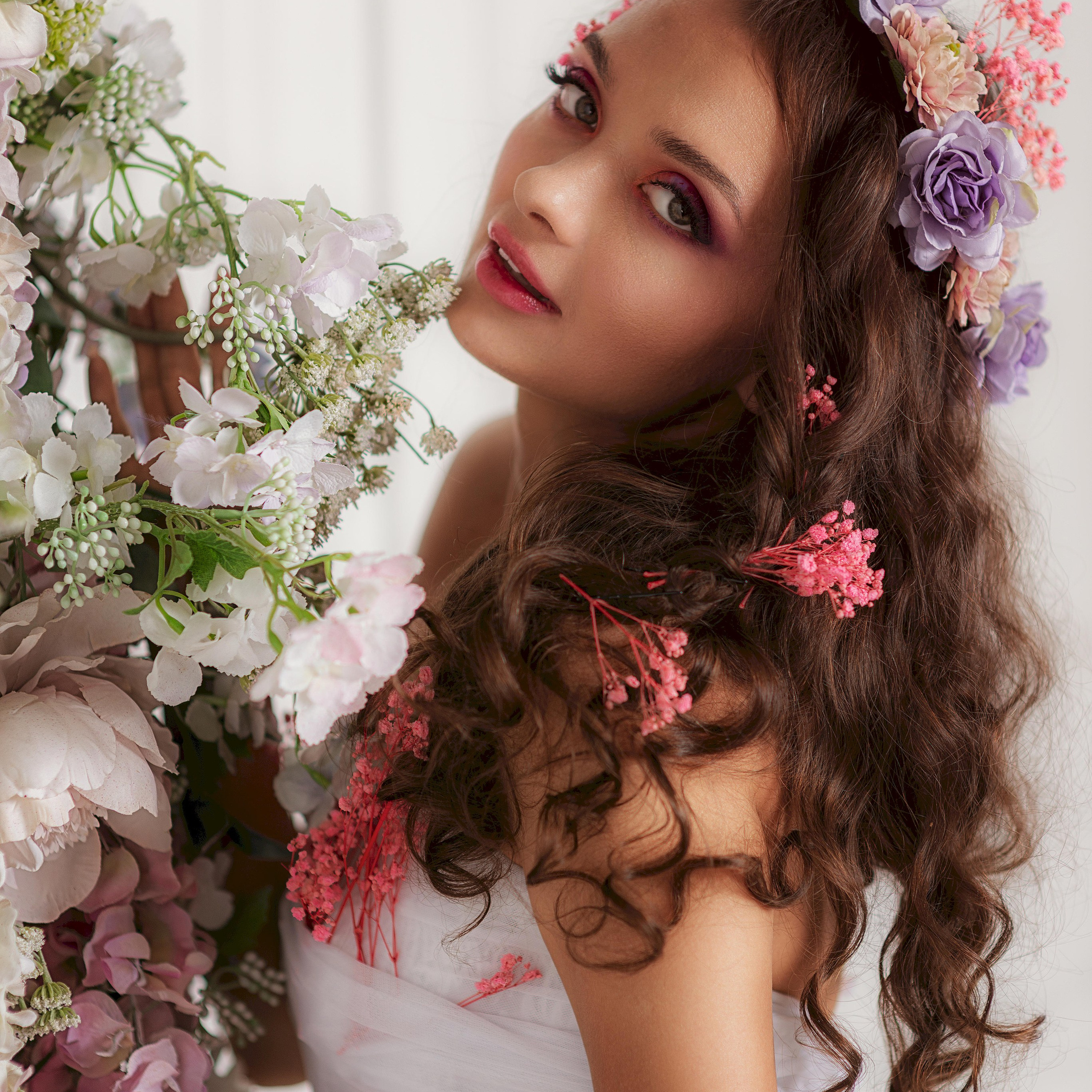 молодая женщина в изысканном образе. Она одета в белое платье с кружевной отделкой, которое подчёркивает нежность и романтичность образа. Её длинные волнистые волосы ниспадают на плечи, дополняя естественную красоту. На голове у неё — венок из цветов пастельных оттенков: сиреневых, розовых и кремовых. В руках она держит пышный букет из живых цветов, который гармонично сочетается с венком.Макияж модели выполнен в насыщенных, но мягких тонах: акцент сделан на глазах с помощью дымчатых теней, губы окрашены в нежный розовый цвет. Поза модели расслабленная и естественная — она слегка наклонила голову, её взгляд направлен в сторону камеры, создавая ощущение лёгкой задумчивости и очарования. Светлый фон подчёркивает цветочную композицию и образ модели, делая снимок воздушным и лёгким.Заголовок«Цветочная симфония: гармония красоты и природы»Ключевые слова: портретная съёмка, венок из цветов, цветочная композиция, белое платье, романтичный образ, макияж, фотосессия, нежность, пастельные тона, фотограф Сафронов Иван, Москва.