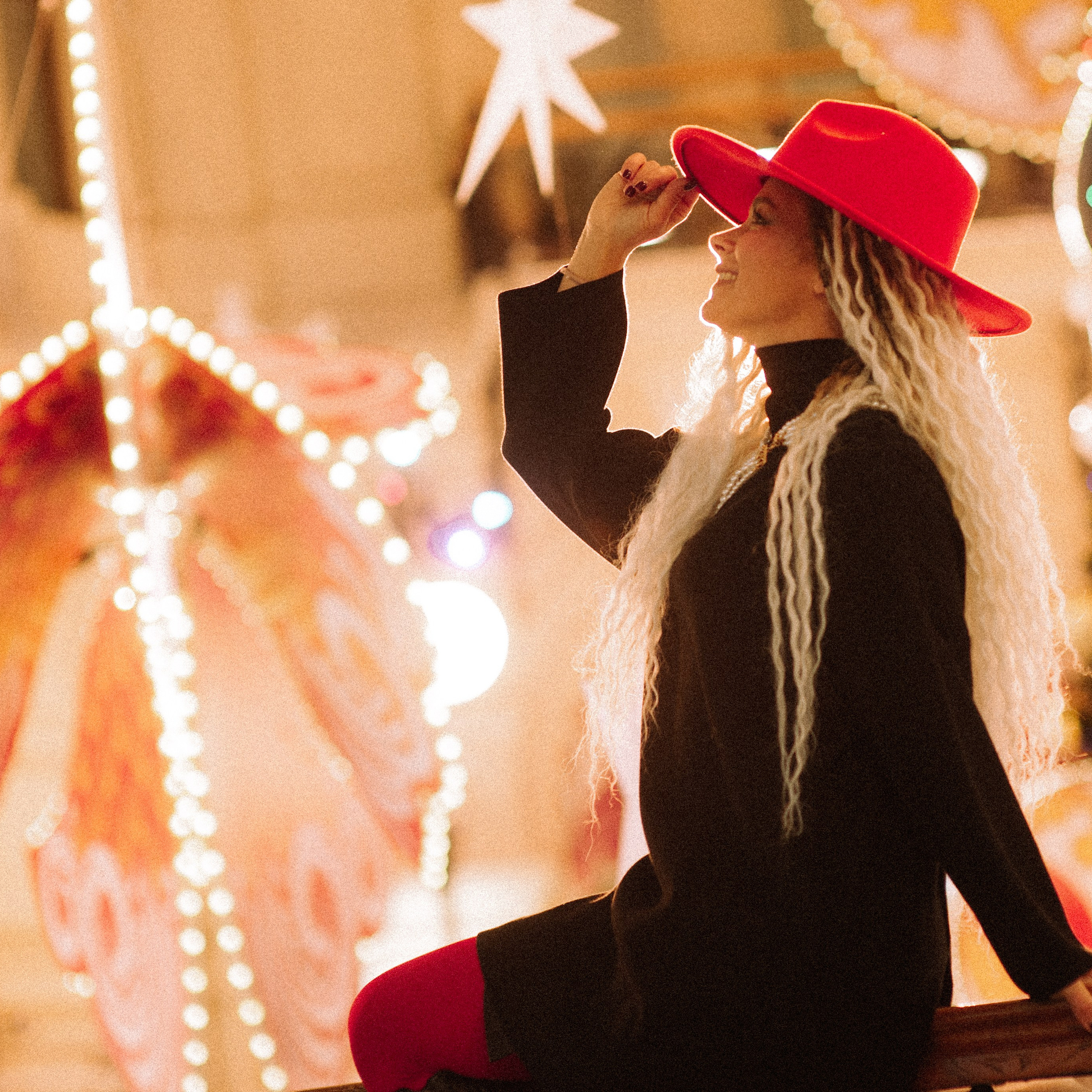 молодая женщина с длинными светлыми дредами. Она одета в стильный чёрный наряд и ярко-красную шляпу, которая сразу привлекает внимание. Девушка сидит, опираясь одной рукой на перила, а другой слегка приподнимает шляпу, создавая игривый и элегантный образ. Её поза расслабленная, но в то же время выразительная — она смотрит вверх, будто наслаждаясь моментом.Фон фотографии наполнен праздничной атмосферой: мерцают огоньки гирлянд, видны элементы новогоднего декора — украшенная ёлка, большие декоративные звёзды и светящиеся фигуры. Тёплый свет гирлянд создаёт уютное, волшебное настроение, подчёркивая контраст между тёмной одеждой модели и яркими акцентами вокруг. Композиция кадра удачно сочетает модель и праздничный фон, делая снимок одновременно стильным и атмосферным.Заголовок«Магия зимних огней: стиль и настроение в одном кадре»Ключевые слова: новогодняя фотосессия, праздничная атмосфера, стильная блондинка, красная шляпа, зимний образ, портретный фотограф в Москве, фотограф Сафронов Иван, праздничные огни, модный лук.