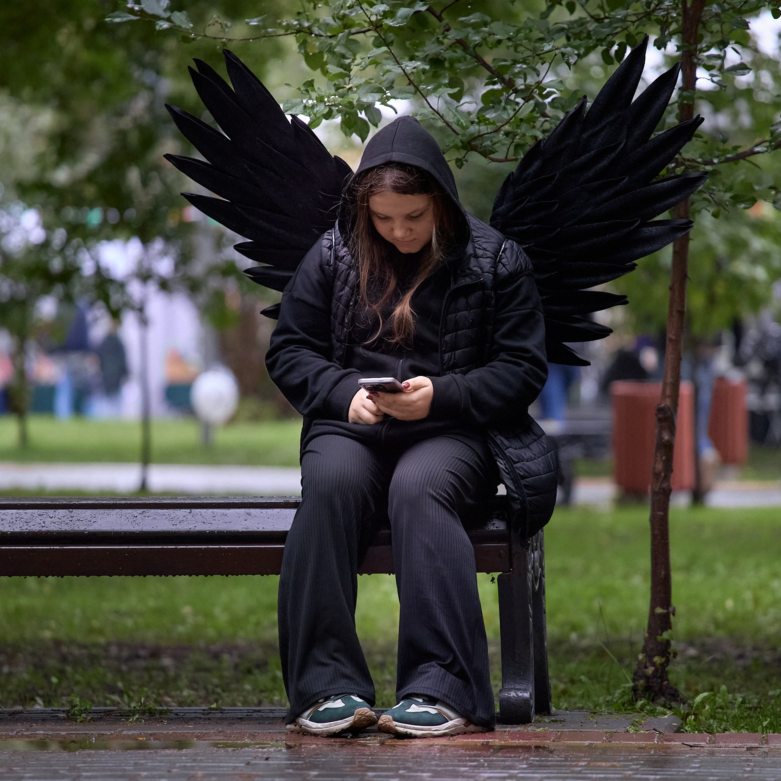 Девочка в костюме черного ангела сидит на лавочке и смотрит в телефон