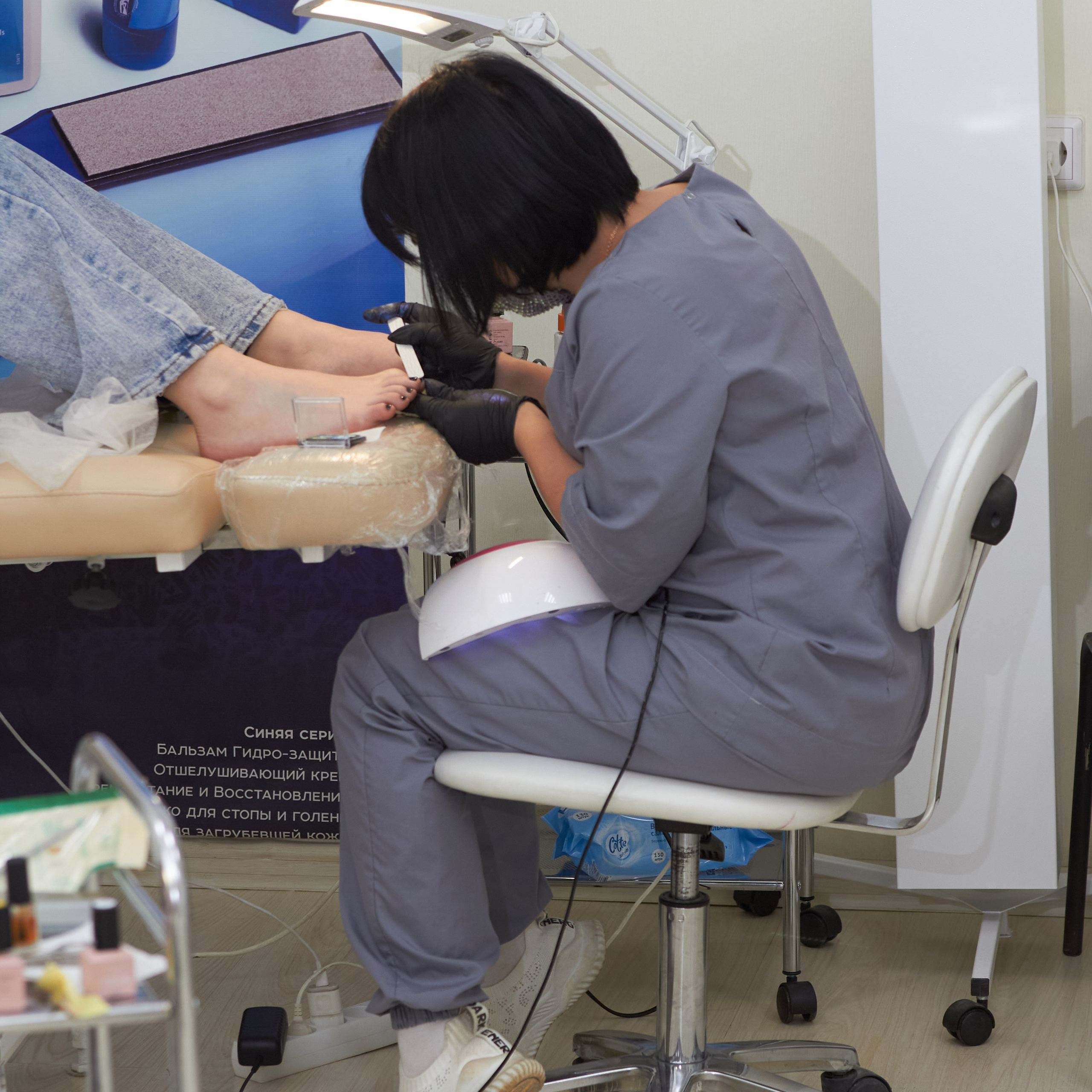 Мастер педикюра в серой униформе склонилась над ножкой клиентки