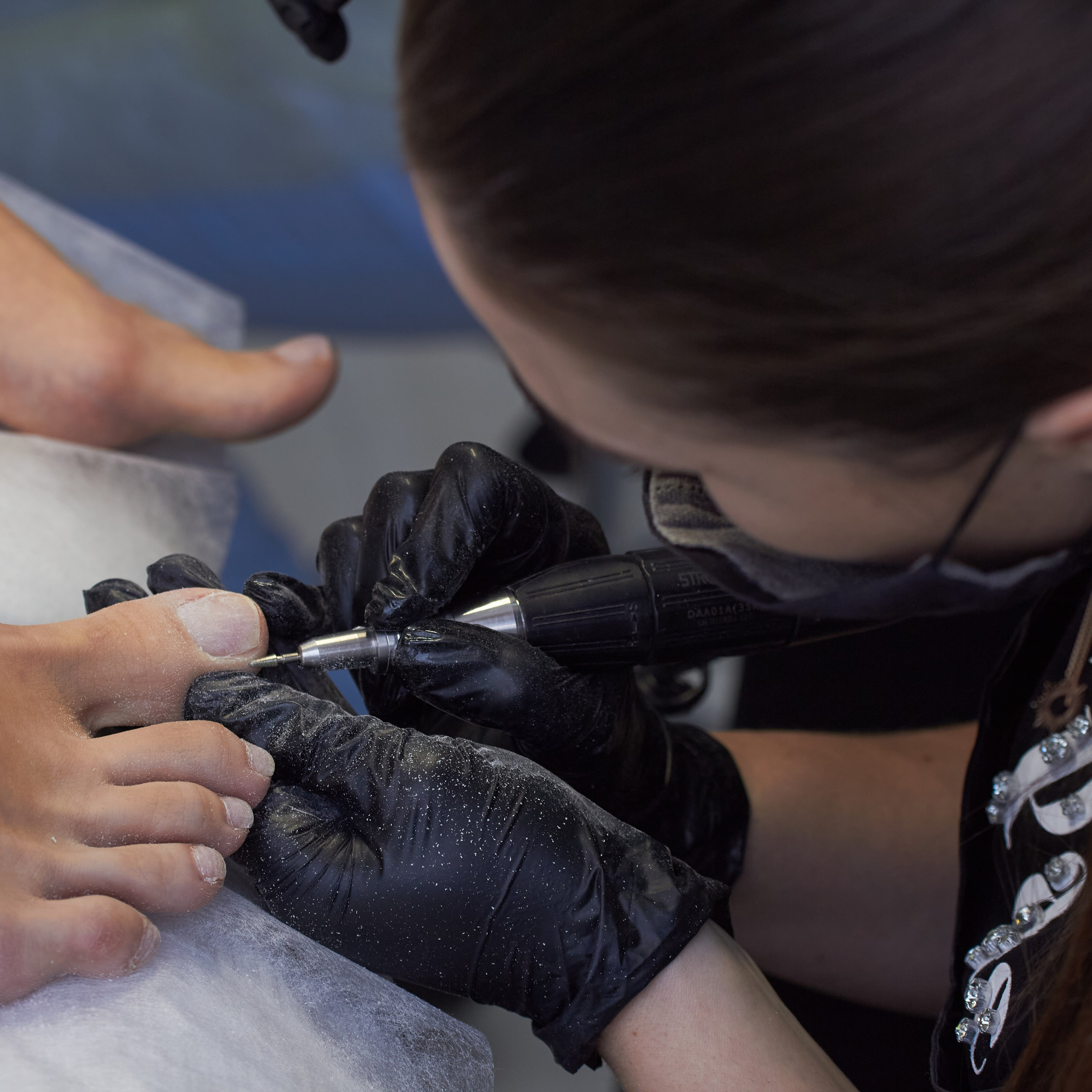 Специалист по педикюру в черной маске и перчатках обтачивает ногти клиенту