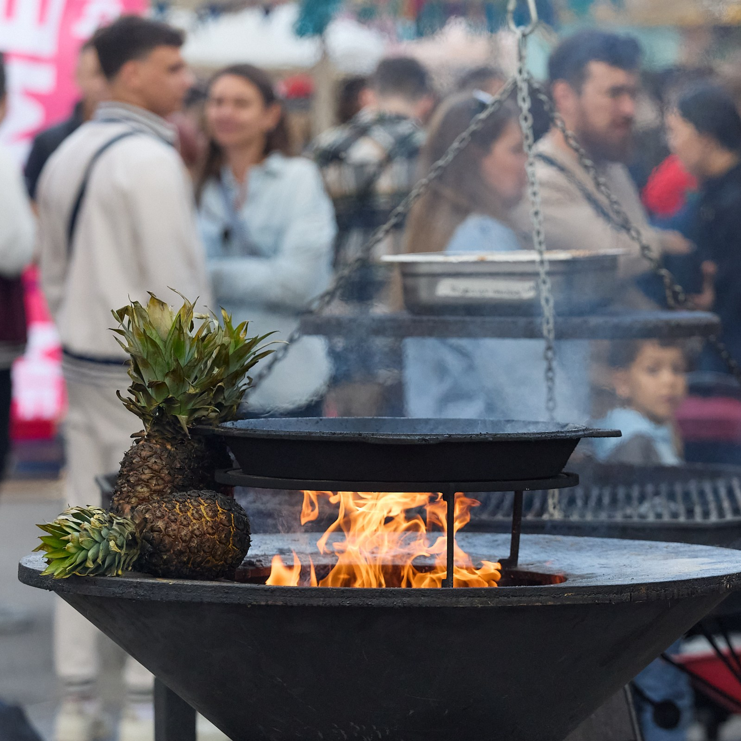 Горит жаровня, сбоку лежат ананасы, позади толпа людей, фото для фуд-фестиваля.