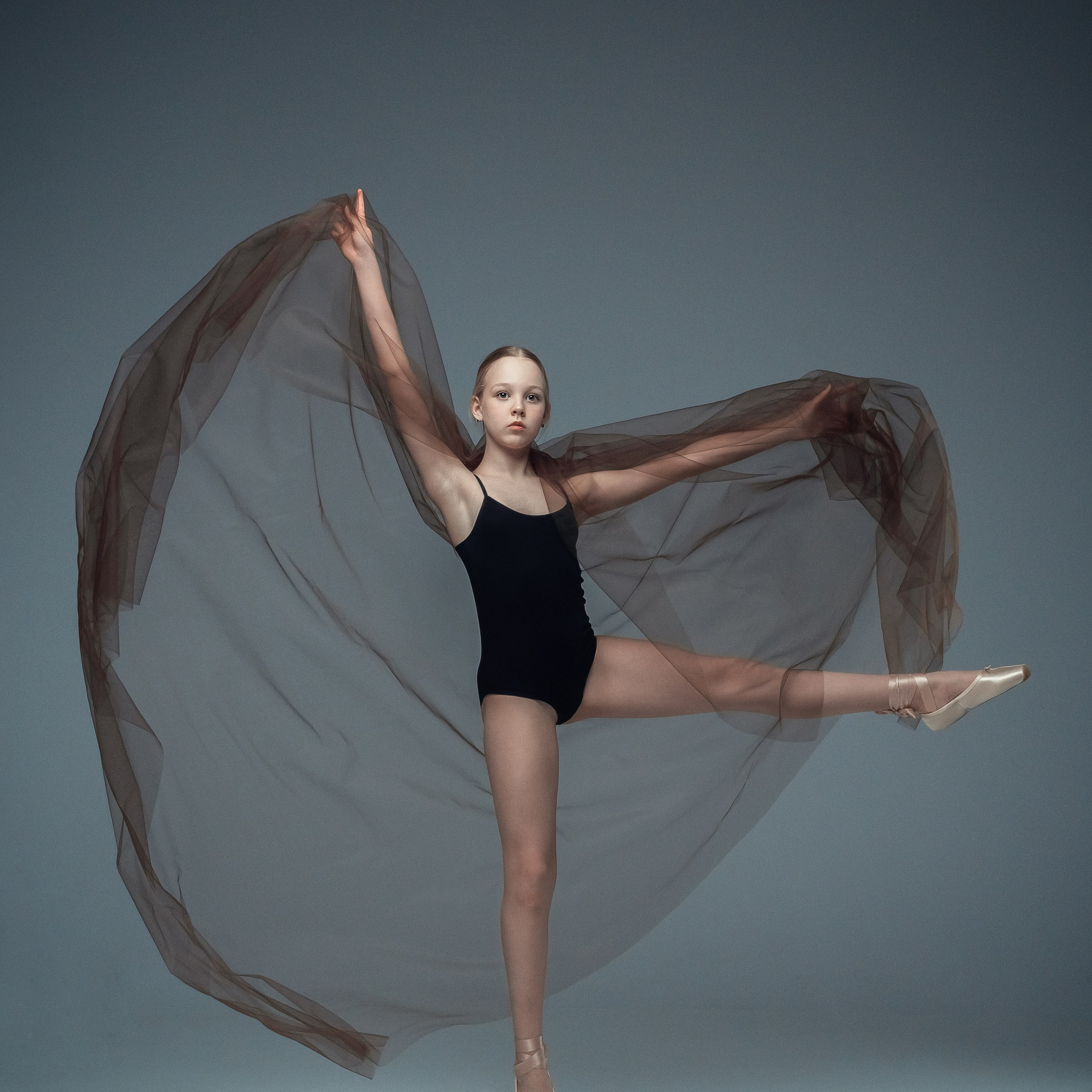 Гимнастка в элегантной позе выполняет элемент с лентой, плавно вытягивая ткань в динамичном движении, демонстрируя гибкость, грацию и выразительность художественной гимнастики