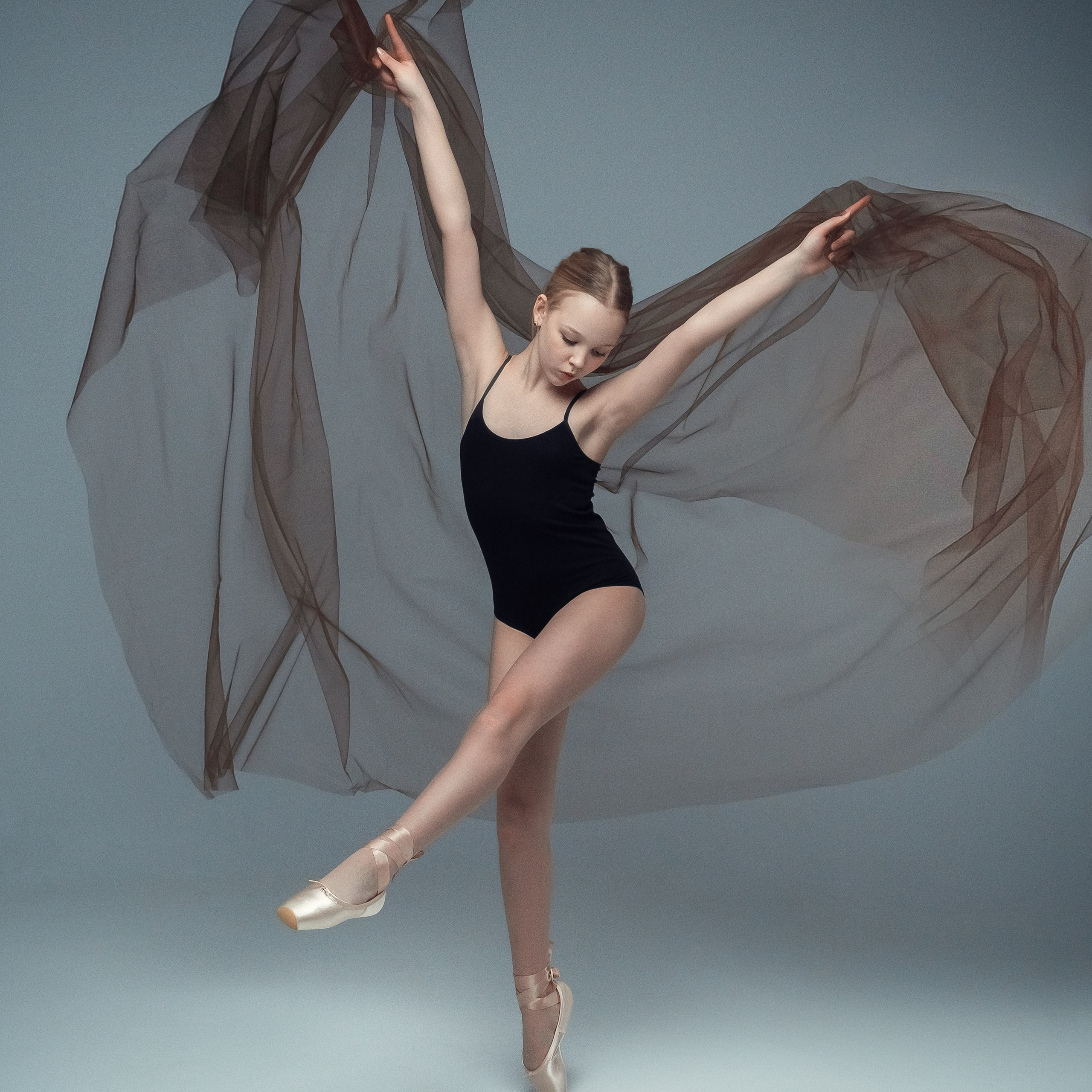 Гимнастка в элегантной позе выполняет элемент с лентой, плавно вытягивая ткань в динамичном движении, демонстрируя гибкость, грацию и выразительность художественной гимнастики