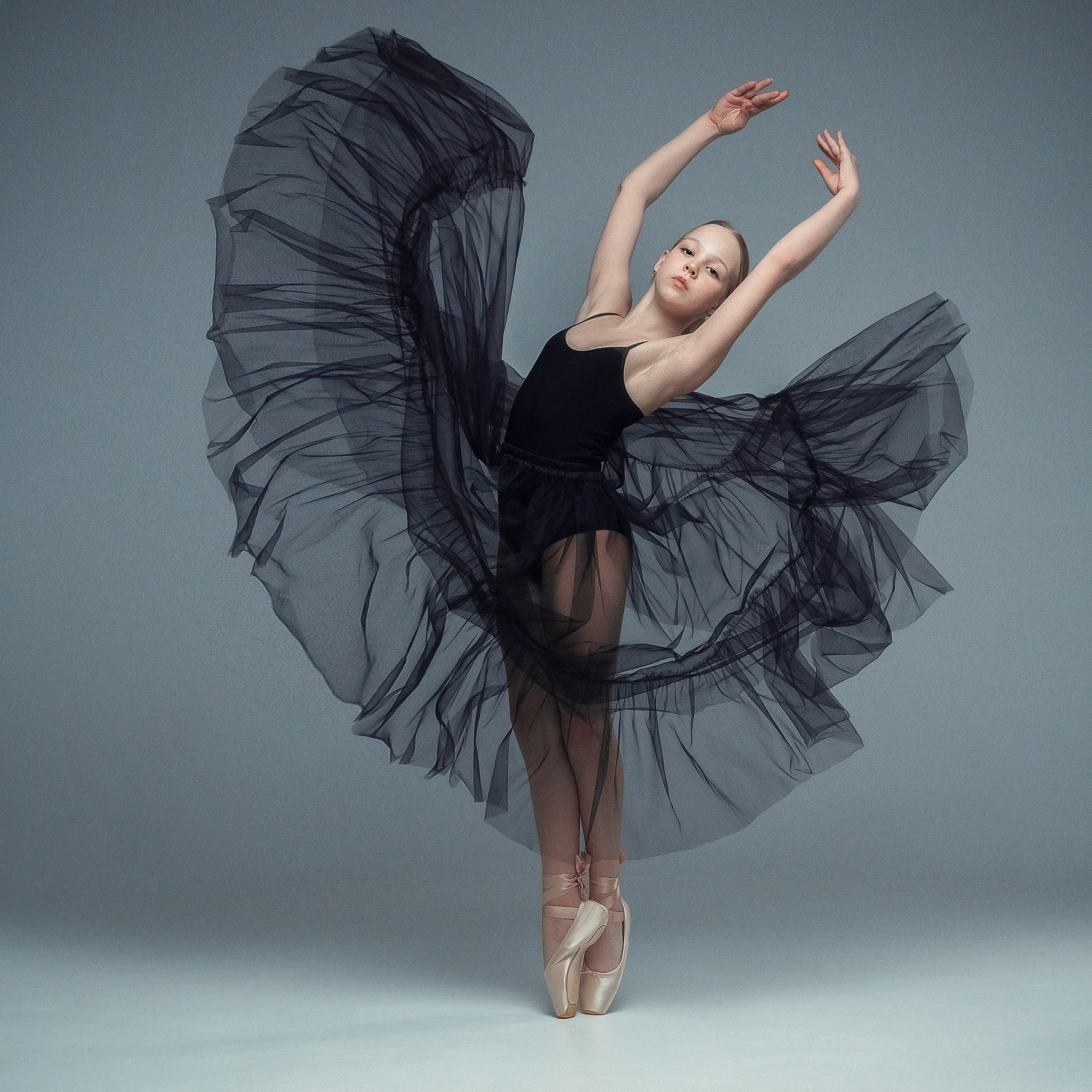 Гимнастка в элегантной позе выполняет элемент с лентой, плавно вытягивая ткань в динамичном движении, демонстрируя гибкость, грацию и выразительность художественной гимнастики