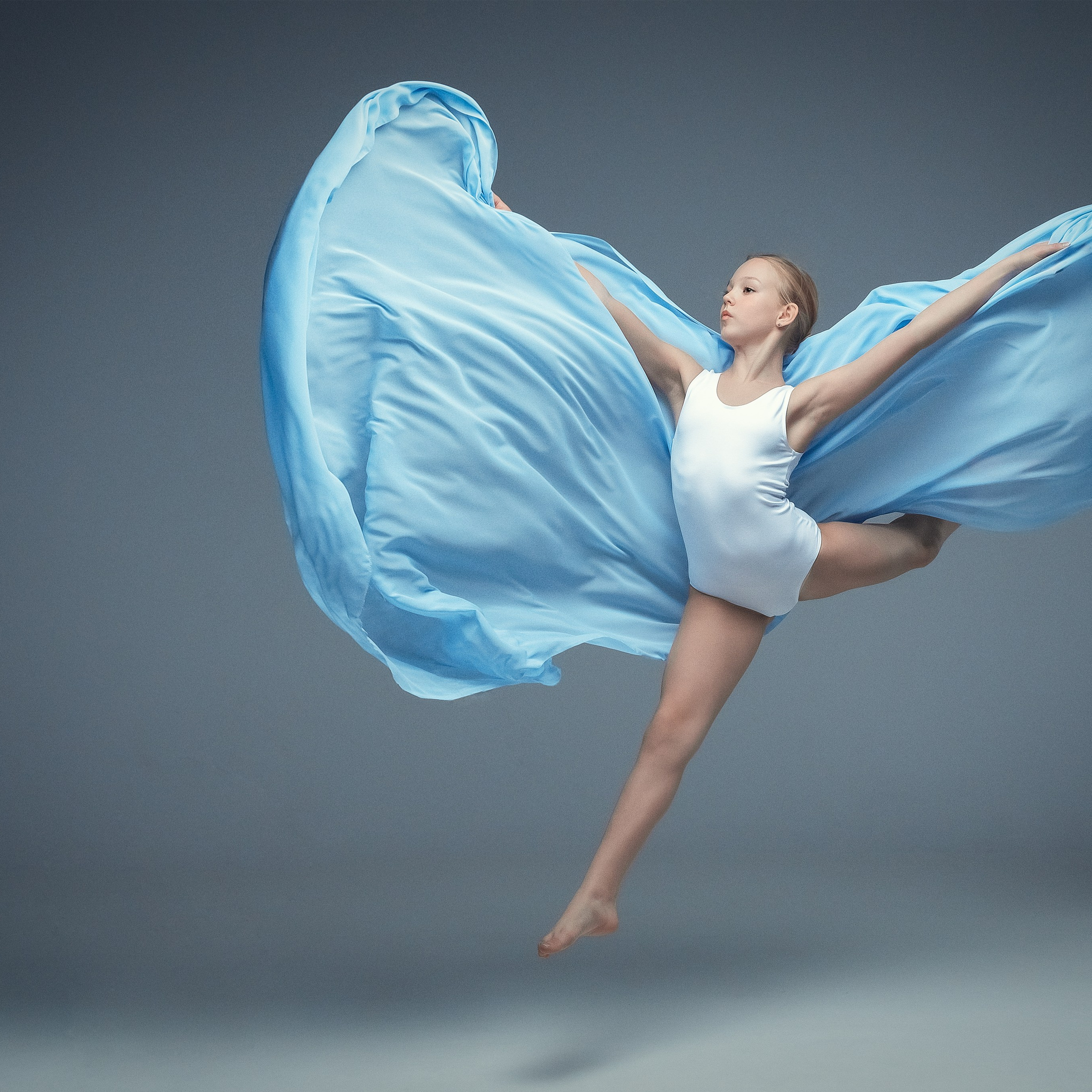 Гимнастка в элегантной позе выполняет элемент с лентой, плавно вытягивая ткань в динамичном движении, демонстрируя гибкость, грацию и выразительность художественной гимнастики