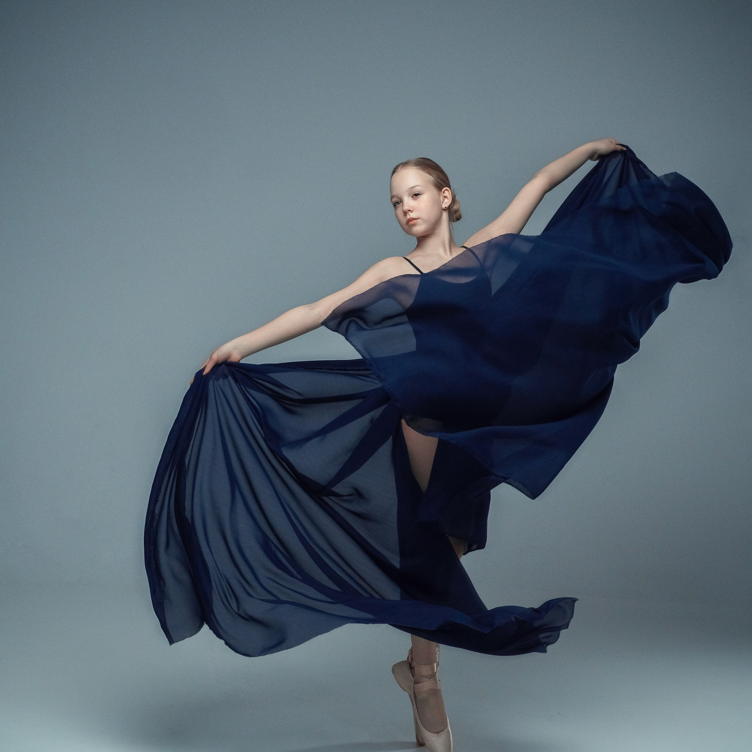 Гимнастка в элегантной позе выполняет элемент с лентой, плавно вытягивая ткань в динамичном движении, демонстрируя гибкость, грацию и выразительность художественной гимнастики