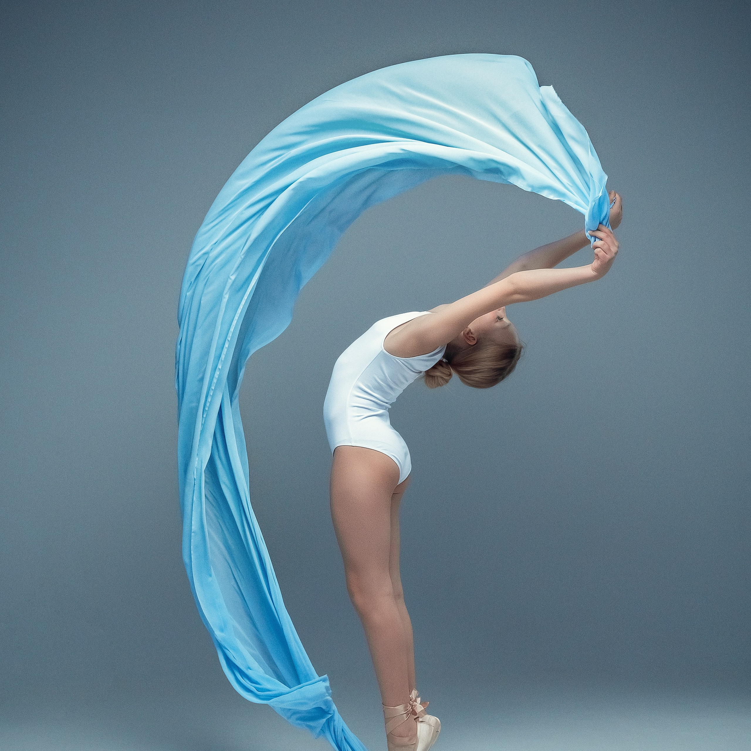 Гимнастка в элегантной позе выполняет элемент с лентой, плавно вытягивая ткань в динамичном движении, демонстрируя гибкость, грацию и выразительность художественной гимнастики