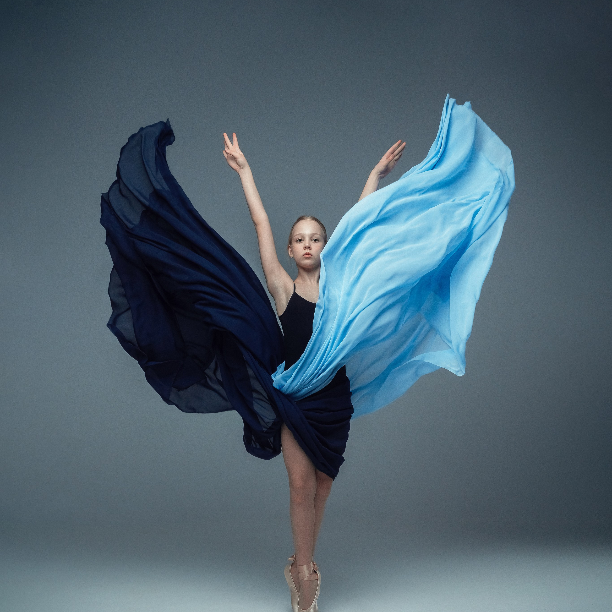 Гимнастка в элегантной позе выполняет элемент с лентой, плавно вытягивая ткань в динамичном движении, демонстрируя гибкость, грацию и выразительность художественной гимнастики