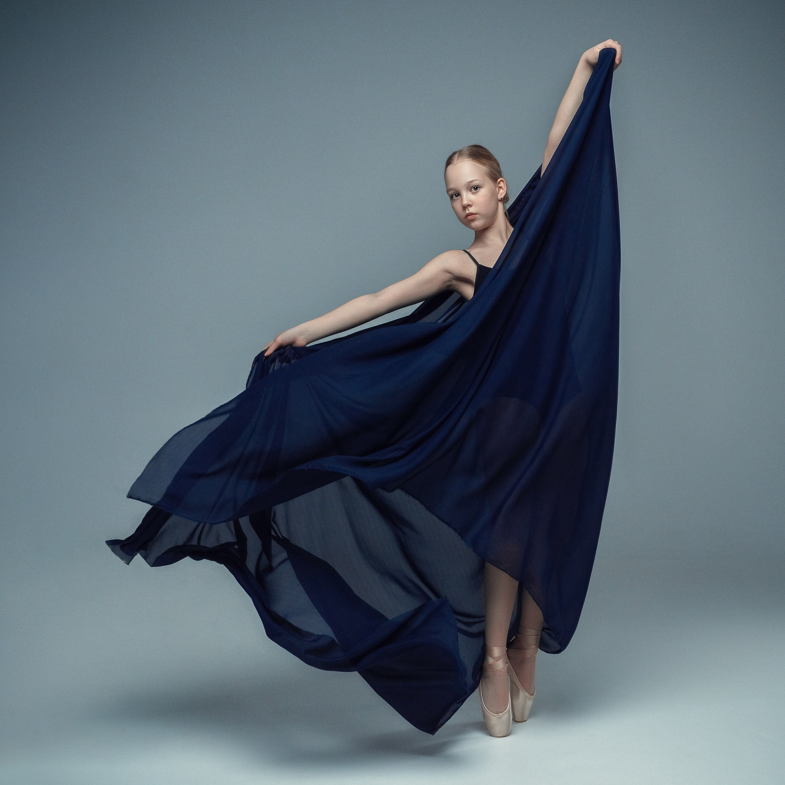 Гимнастка в элегантной позе выполняет элемент с лентой, плавно вытягивая ткань в динамичном движении, демонстрируя гибкость, грацию и выразительность художественной гимнастики