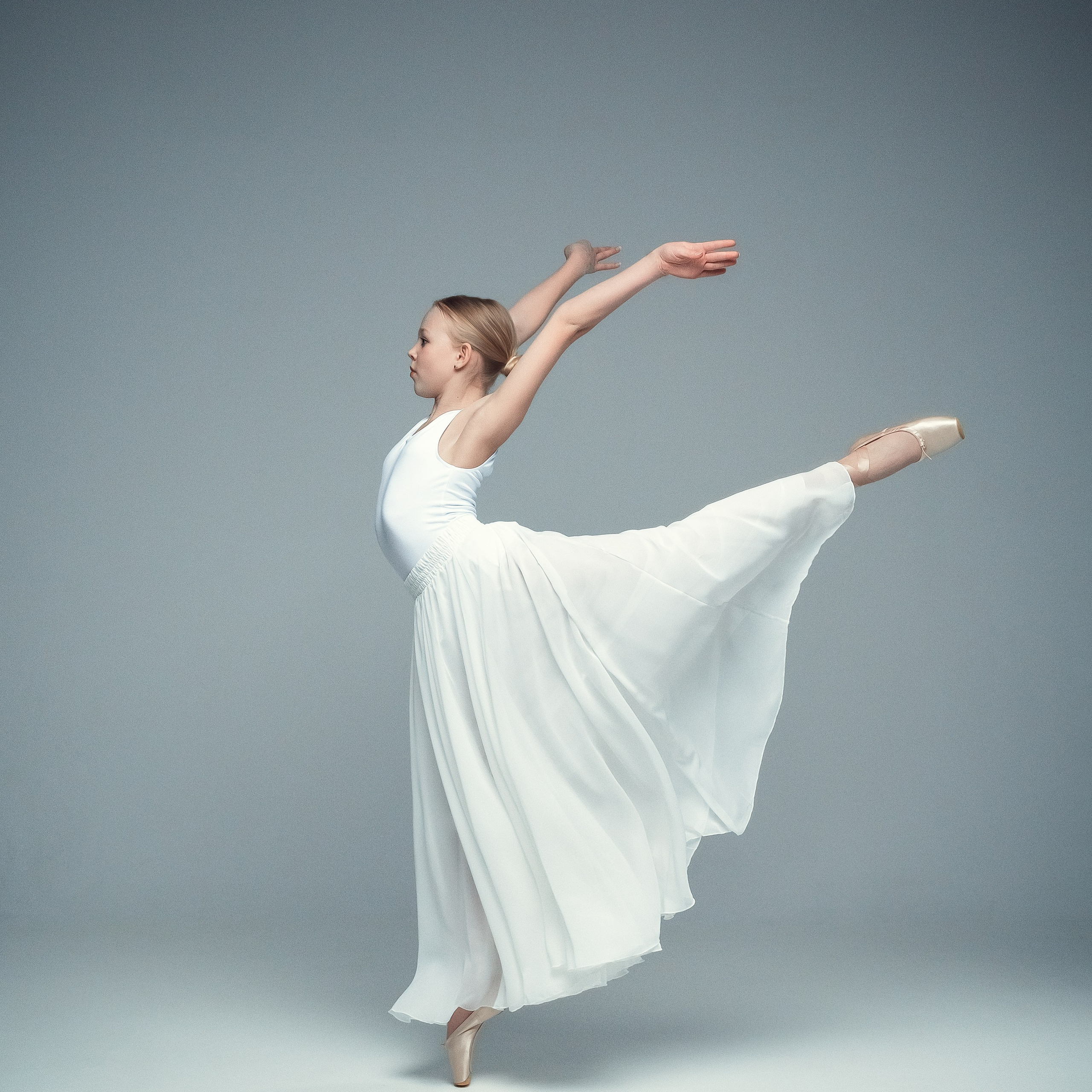 Гимнастка в элегантной позе выполняет элемент с лентой, плавно вытягивая ткань в динамичном движении, демонстрируя гибкость, грацию и выразительность художественной гимнастики