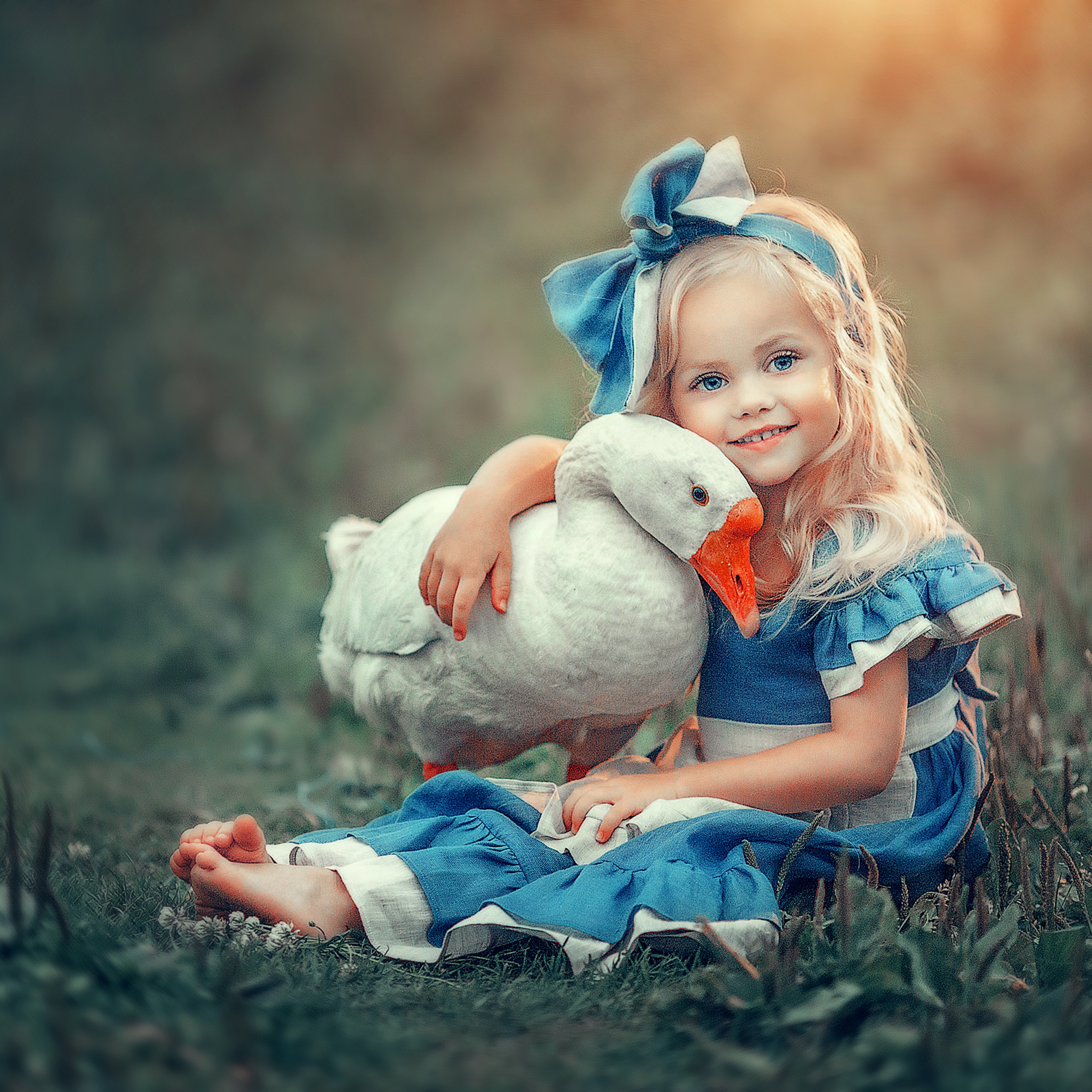 Красивая девочка со светлыми волосами в голубом платье  сидит на траве с гусем